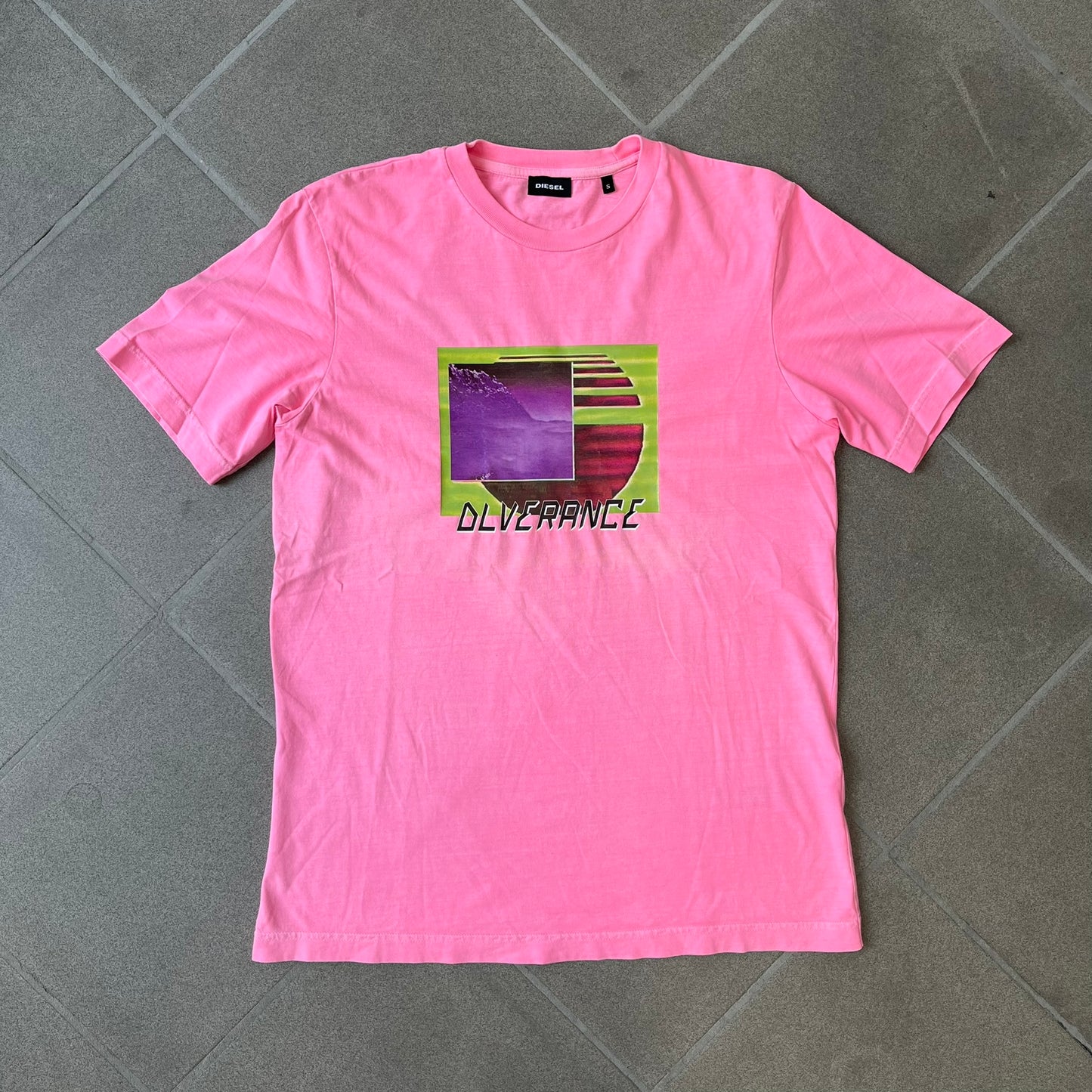 Diesel DLVERANCE Bright Pink Tee features a 100% cotton jersey construction with a fluorescent pink pigment-dyed finish. The front has a rubberised “DLVERANCE” graphic, with a small rubber “Copyright” logo on the lower back.
