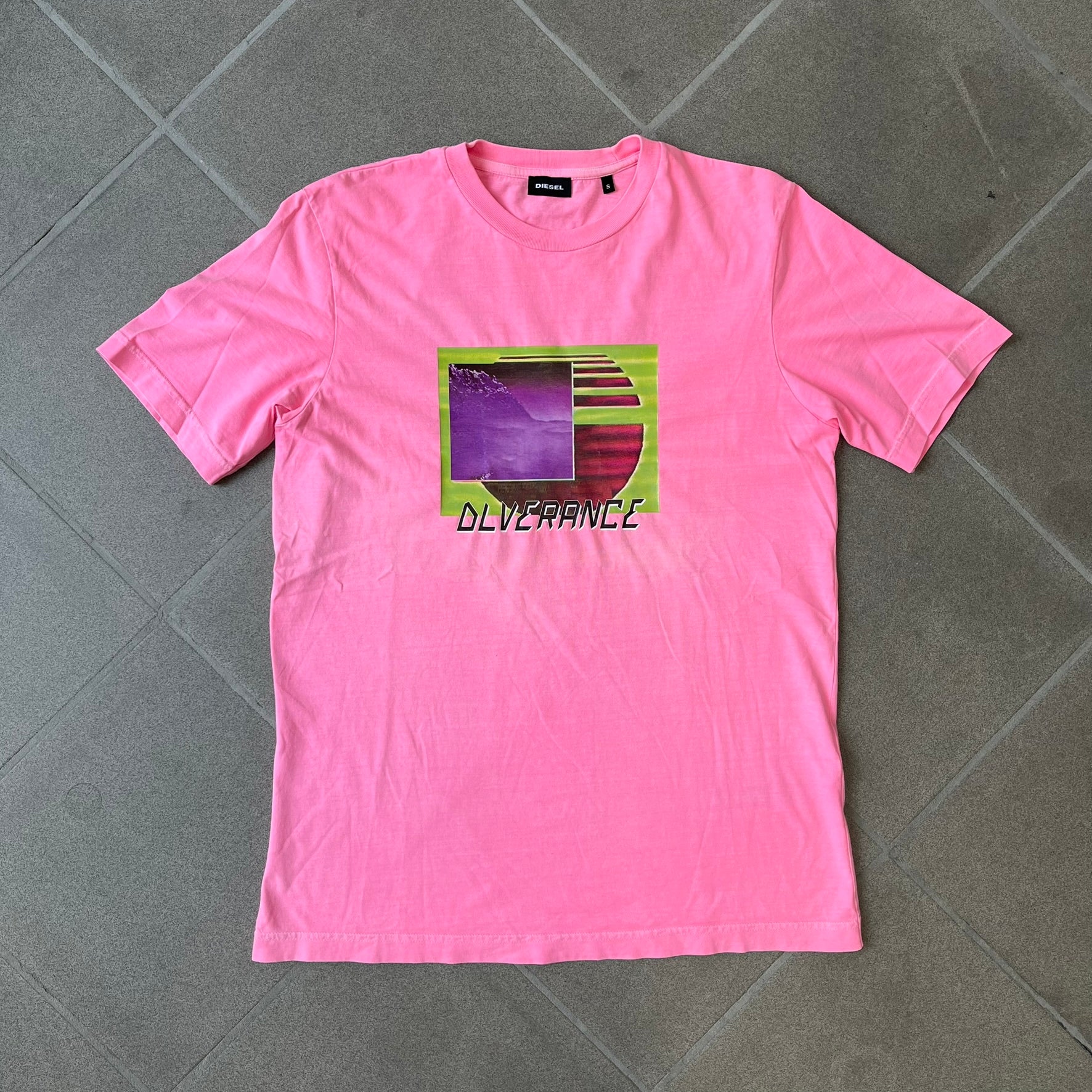 Diesel DLVERANCE Bright Pink Tee features a 100% cotton jersey construction with a fluorescent pink pigment-dyed finish. The front has a rubberised “DLVERANCE” graphic, with a small rubber “Copyright” logo on the lower back.