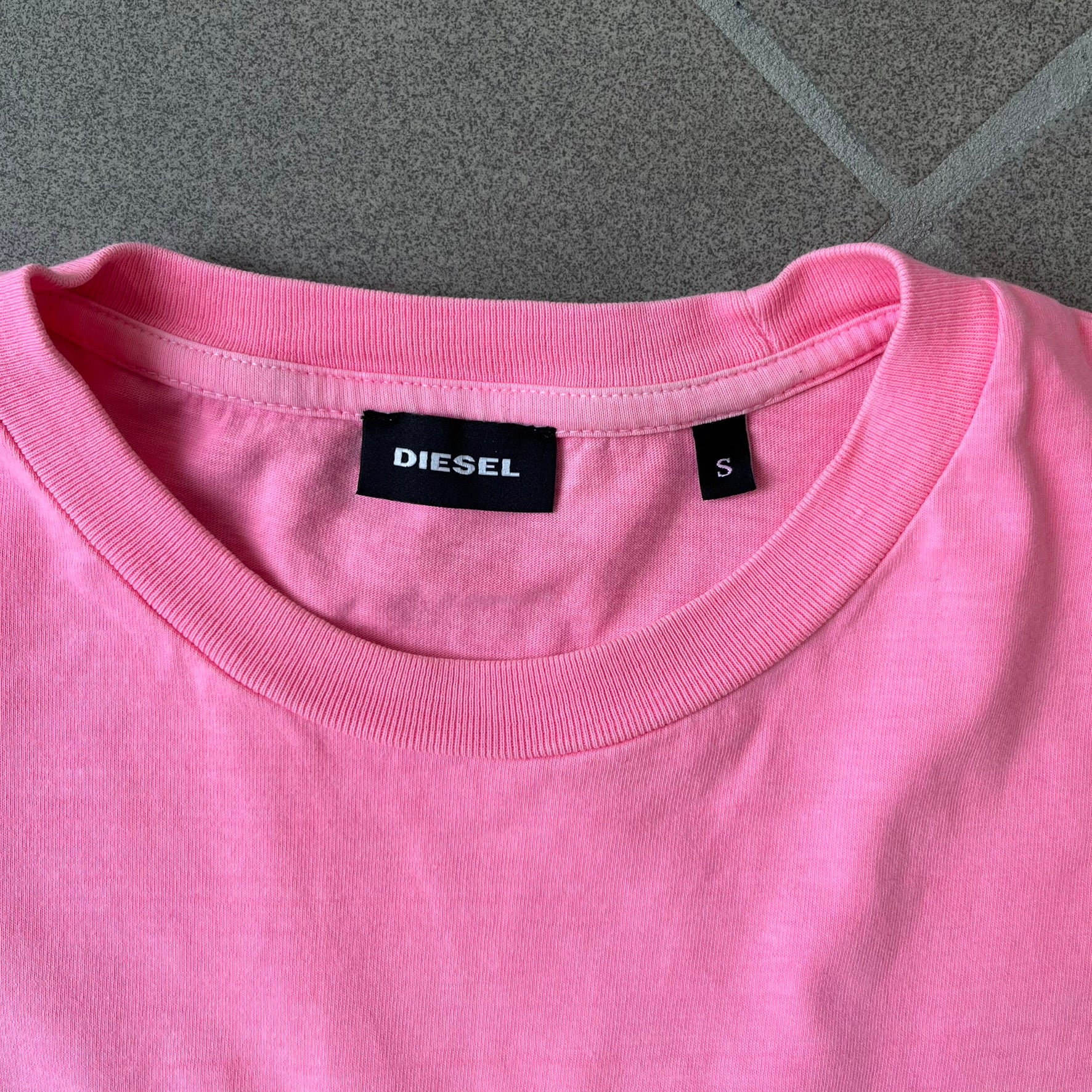Diesel DLVERANCE Bright Pink Tee features a 100% cotton jersey construction with a fluorescent pink pigment-dyed finish. The front has a rubberised “DLVERANCE” graphic, with a small rubber “Copyright” logo on the lower back.
