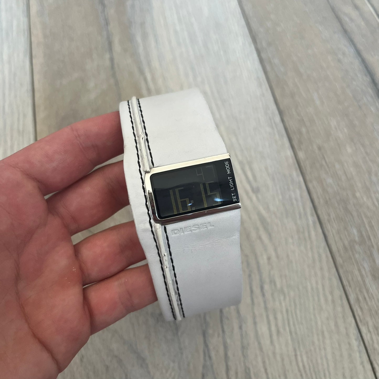 Diesel DZ7054 White Leather Strap Digital Watch features a White leather strap and a black digital display housed in a silver rectangular case. The watch includes a backlight, Arabic numeral indices, and a deployant closure, with a stainless steel case back engraved with Diesel branding and model number DZ-7090. It is water resistant up to 50 meters (5 ATM) and combines industrial design with functional quartz digital movement.&nbsp;