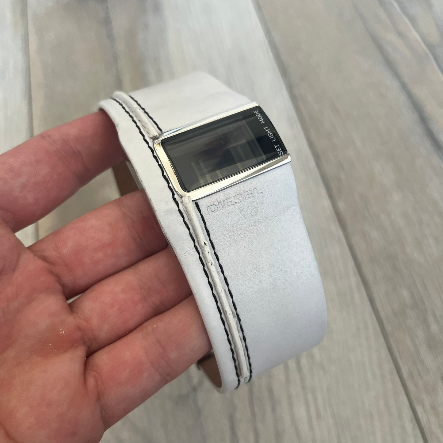 Diesel DZ7054 White Leather Strap Digital Watch features a White leather strap and a black digital display housed in a silver rectangular case. The watch includes a backlight, Arabic numeral indices, and a deployant closure, with a stainless steel case back engraved with Diesel branding and model number DZ-7090. It is water resistant up to 50 meters (5 ATM) and combines industrial design with functional quartz digital movement.&nbsp;