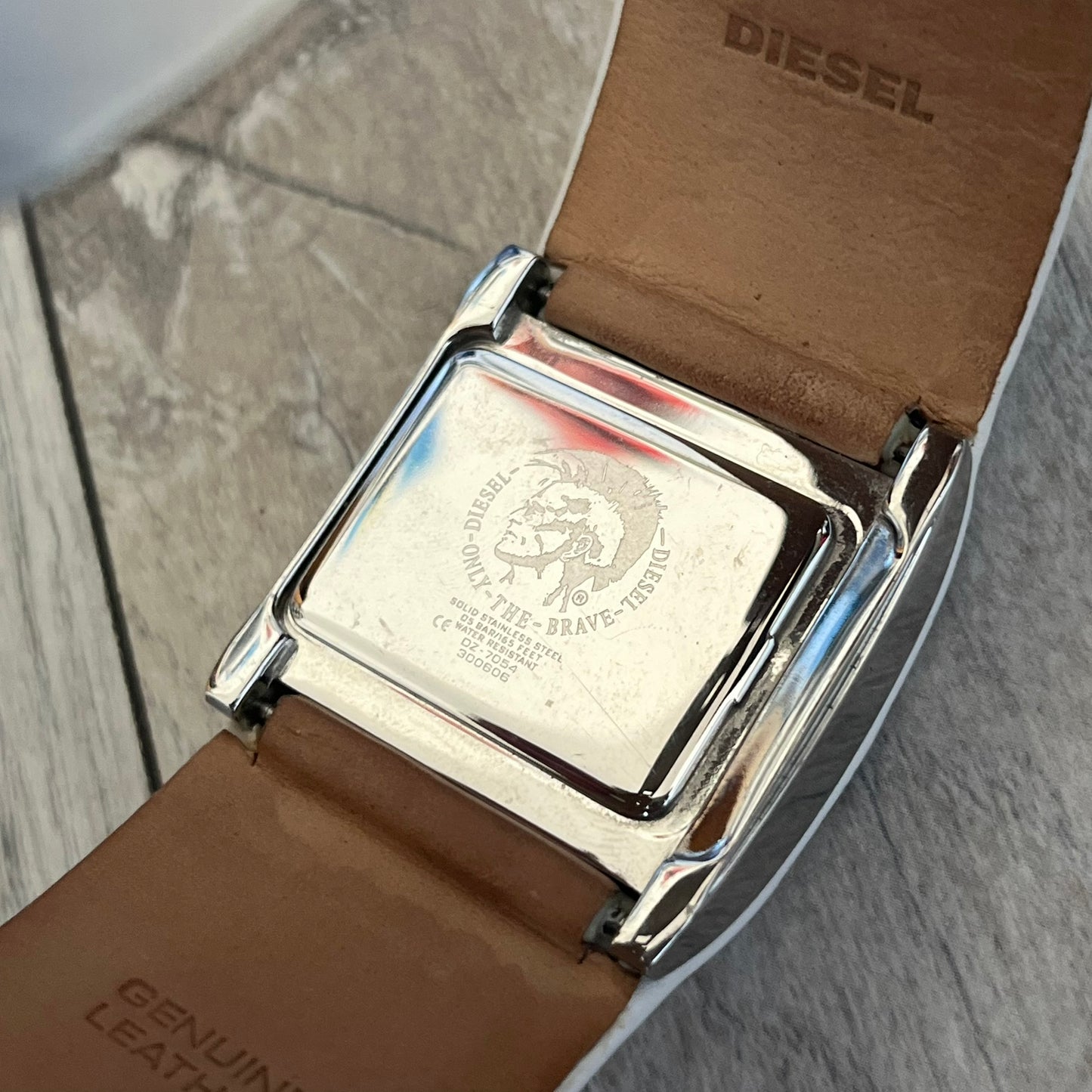 Diesel DZ7054 White Leather Strap Digital Watch features a White leather strap and a black digital display housed in a silver rectangular case. The watch includes a backlight, Arabic numeral indices, and a deployant closure, with a stainless steel case back engraved with Diesel branding and model number DZ-7090. It is water resistant up to 50 meters (5 ATM) and combines industrial design with functional quartz digital movement.&nbsp;