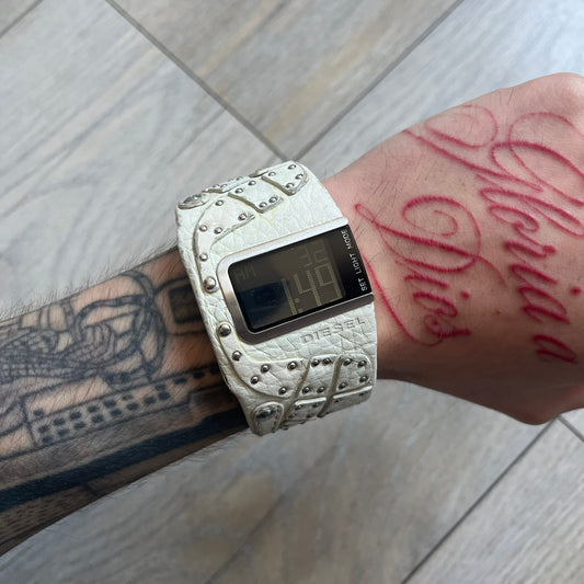 Diesel DZ7068 Studded Leather Strap Digital Watch features a white leather patchwork strap accented with small metal studs and a black digital display housed in a silver rectangular case. The watch includes a backlight, Arabic numeral indices, and a deployant closure, with a stainless steel case back engraved with Diesel branding and model number DZ-7068. It is water resistant up to 50 meters (5 ATM) and combines industrial design with functional quartz digital movement.