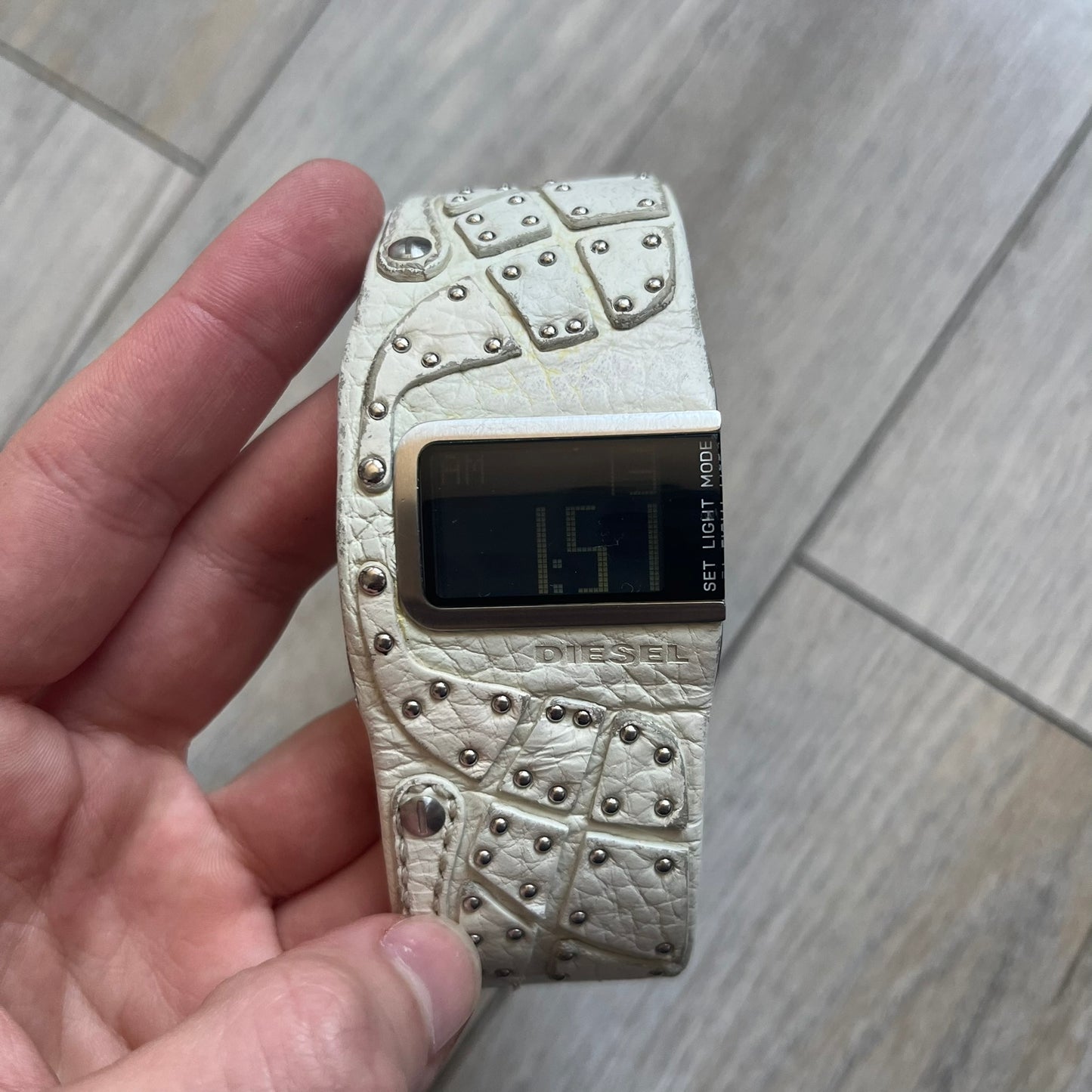 Diesel DZ7068 Studded Leather Strap Digital Watch features a white leather patchwork strap accented with small metal studs and a black digital display housed in a silver rectangular case. The watch includes a backlight, Arabic numeral indices, and a deployant closure, with a stainless steel case back engraved with Diesel branding and model number DZ-7068. It is water resistant up to 50 meters (5 ATM) and combines industrial design with functional quartz digital movement.