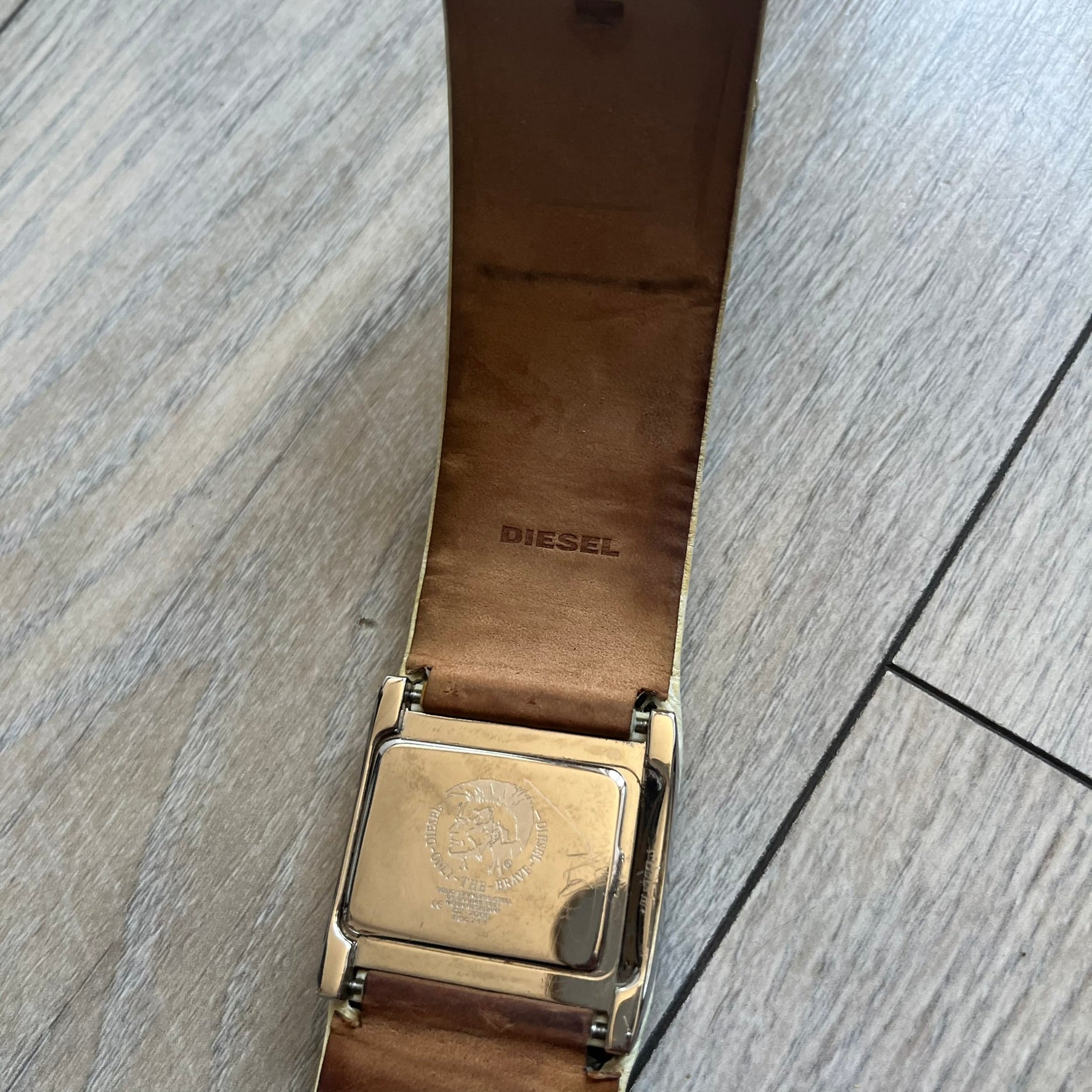 Diesel DZ7068 Studded Leather Strap Digital Watch features a white leather patchwork strap accented with small metal studs and a black digital display housed in a silver rectangular case. The watch includes a backlight, Arabic numeral indices, and a deployant closure, with a stainless steel case back engraved with Diesel branding and model number DZ-7068. It is water resistant up to 50 meters (5 ATM) and combines industrial design with functional quartz digital movement.