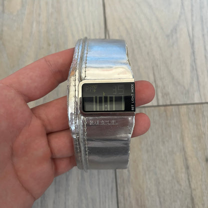 Diesel DZ7090 Silver Leather Strap Digital Watch features a Silver leather patchwork strap and a black digital display housed in a silver rectangular case. The watch includes a backlight, Arabic numeral indices, and a deployant closure, with a stainless steel case back engraved with Diesel branding and model number DZ-7090. It is water resistant up to 50 meters (5 ATM) and combines industrial design with functional quartz digital movement.&nbsp;