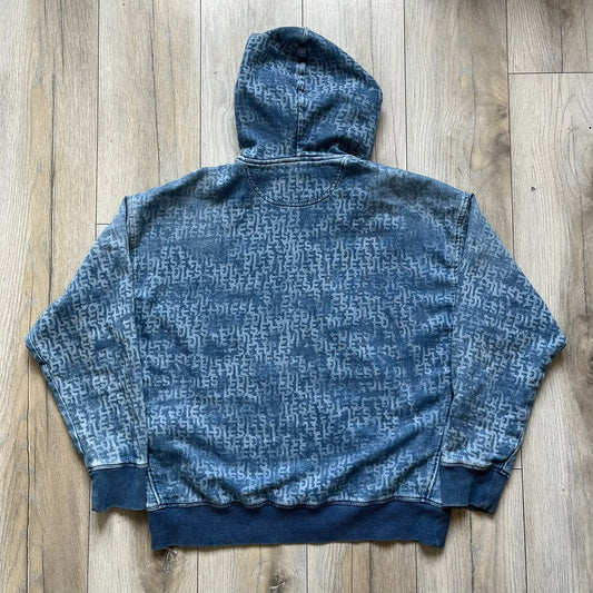 Diesel “Denim” Logo Blue Hoodie is a blue hooded sweatshirt designed with a denim-inspired colorway and washed effect throughout. The fabric features varying light and dark blue shades to mimic the appearance of worn denim, along with an all-over Diesel text pattern in a slightly lighter tone. A small red Diesel patch is placed on the chest, while the cuffs and hem are finished in a contrasting navy color.&nbsp;