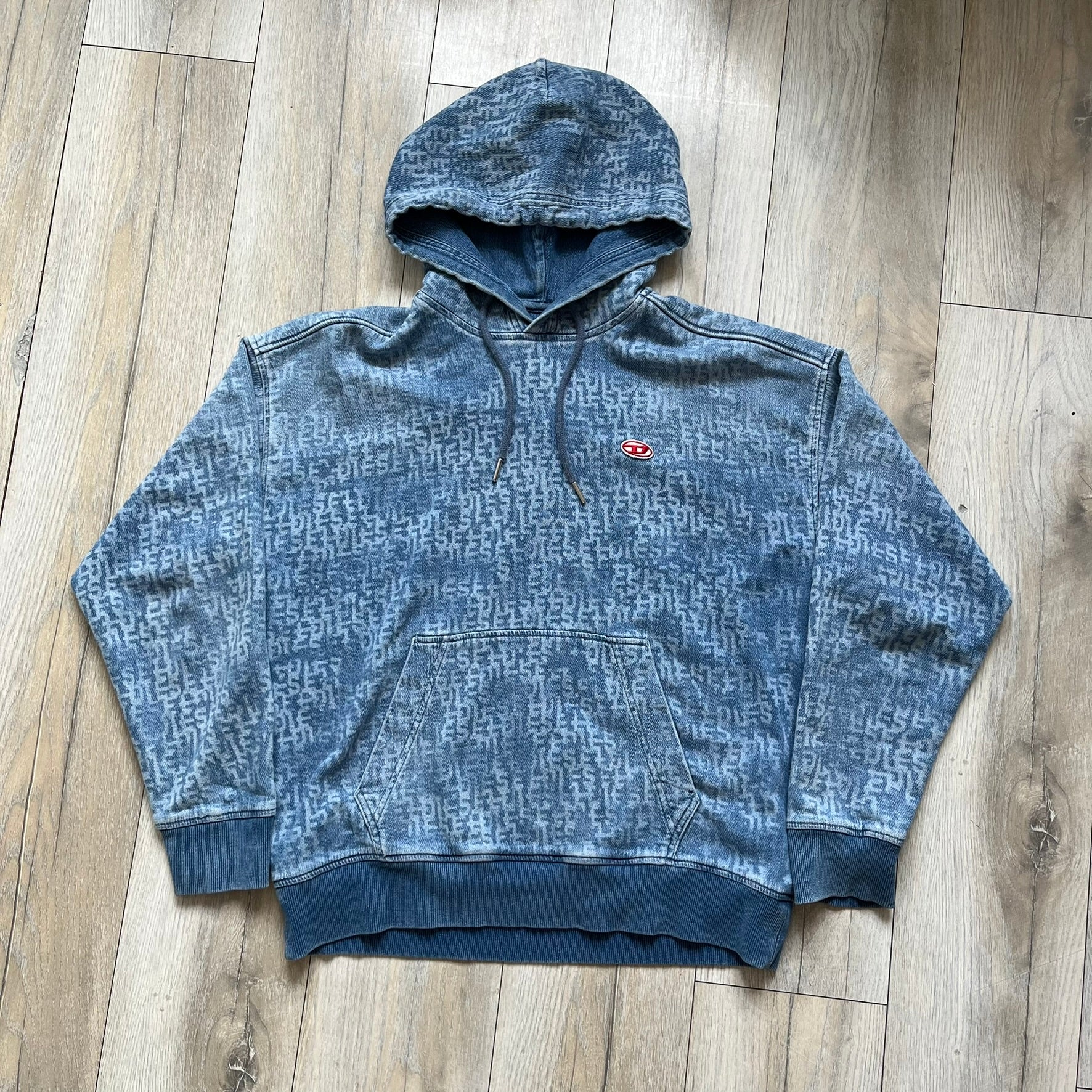 Diesel “Denim” Logo Blue Hoodie is a blue hooded sweatshirt designed with a denim-inspired colorway and washed effect throughout. The fabric features varying light and dark blue shades to mimic the appearance of worn denim, along with an all-over Diesel text pattern in a slightly lighter tone. A small red Diesel patch is placed on the chest, while the cuffs and hem are finished in a contrasting navy color.&nbsp;