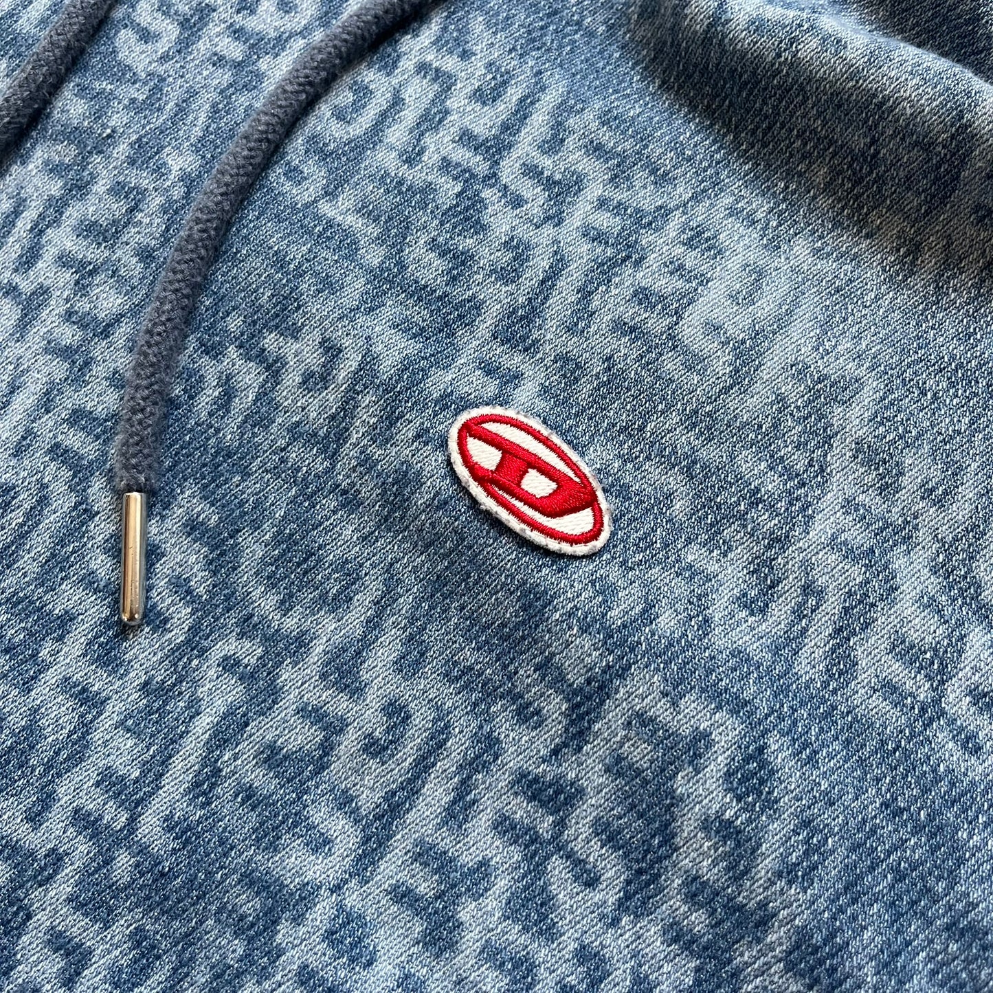 Diesel “Denim” Logo Blue Hoodie is a blue hooded sweatshirt designed with a denim-inspired colorway and washed effect throughout. The fabric features varying light and dark blue shades to mimic the appearance of worn denim, along with an all-over Diesel text pattern in a slightly lighter tone. A small red Diesel patch is placed on the chest, while the cuffs and hem are finished in a contrasting navy color.&nbsp;