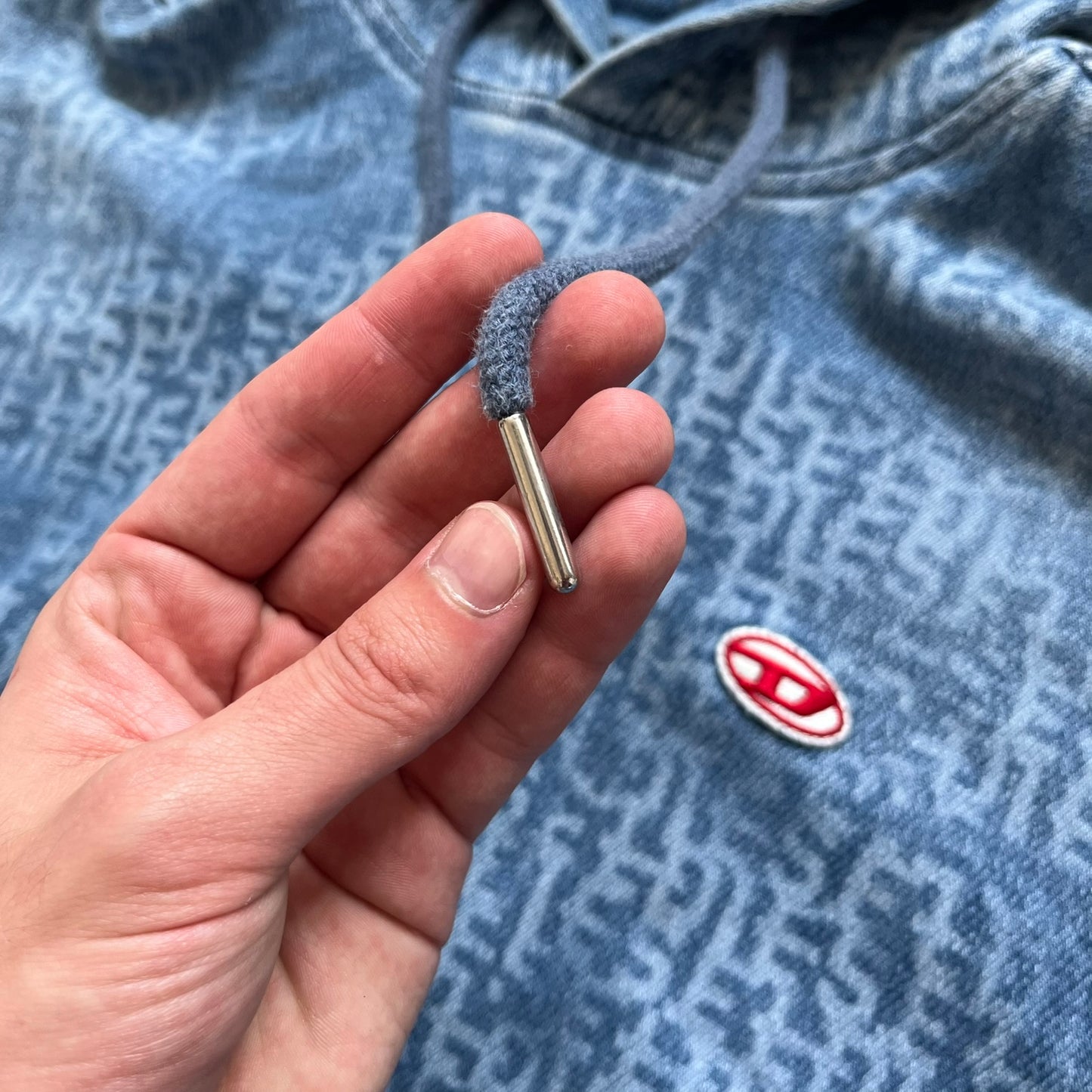 Diesel “Denim” Logo Blue Hoodie is a blue hooded sweatshirt designed with a denim-inspired colorway and washed effect throughout. The fabric features varying light and dark blue shades to mimic the appearance of worn denim, along with an all-over Diesel text pattern in a slightly lighter tone. A small red Diesel patch is placed on the chest, while the cuffs and hem are finished in a contrasting navy color.&nbsp;