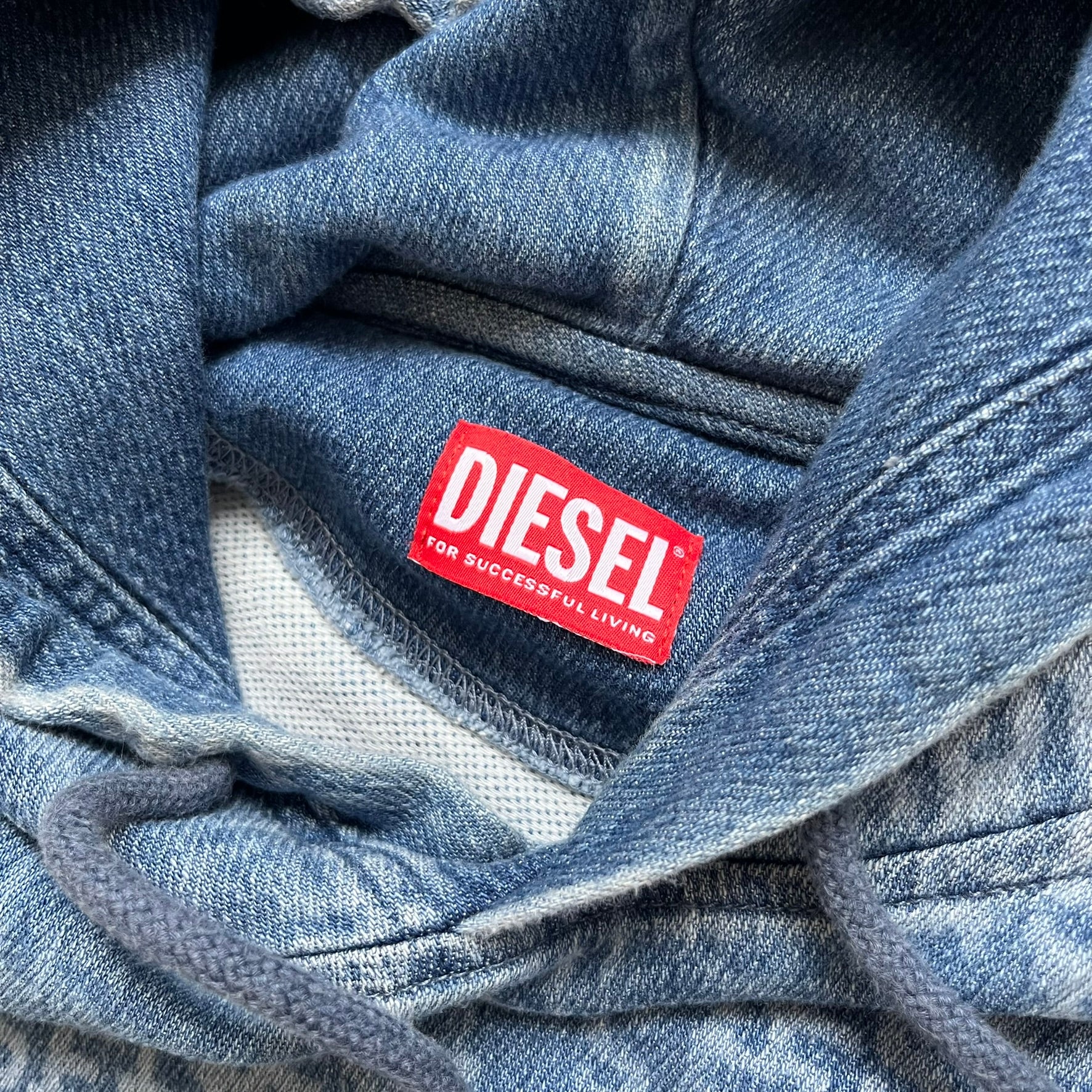 Diesel “Denim” Logo Blue Hoodie is a blue hooded sweatshirt designed with a denim-inspired colorway and washed effect throughout. The fabric features varying light and dark blue shades to mimic the appearance of worn denim, along with an all-over Diesel text pattern in a slightly lighter tone. A small red Diesel patch is placed on the chest, while the cuffs and hem are finished in a contrasting navy color.&nbsp;