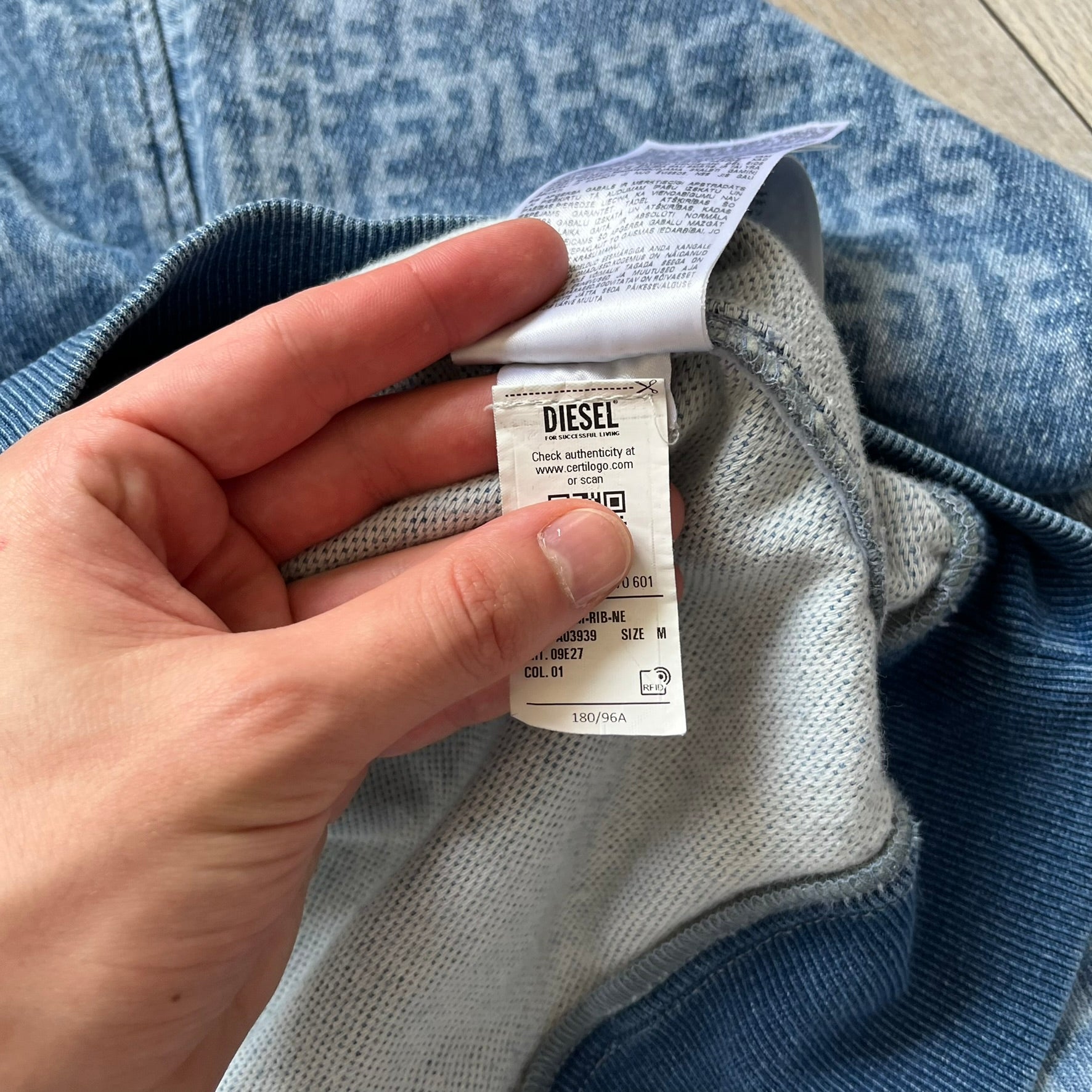 Diesel “Denim” Logo Blue Hoodie is a blue hooded sweatshirt designed with a denim-inspired colorway and washed effect throughout. The fabric features varying light and dark blue shades to mimic the appearance of worn denim, along with an all-over Diesel text pattern in a slightly lighter tone. A small red Diesel patch is placed on the chest, while the cuffs and hem are finished in a contrasting navy color.&nbsp;