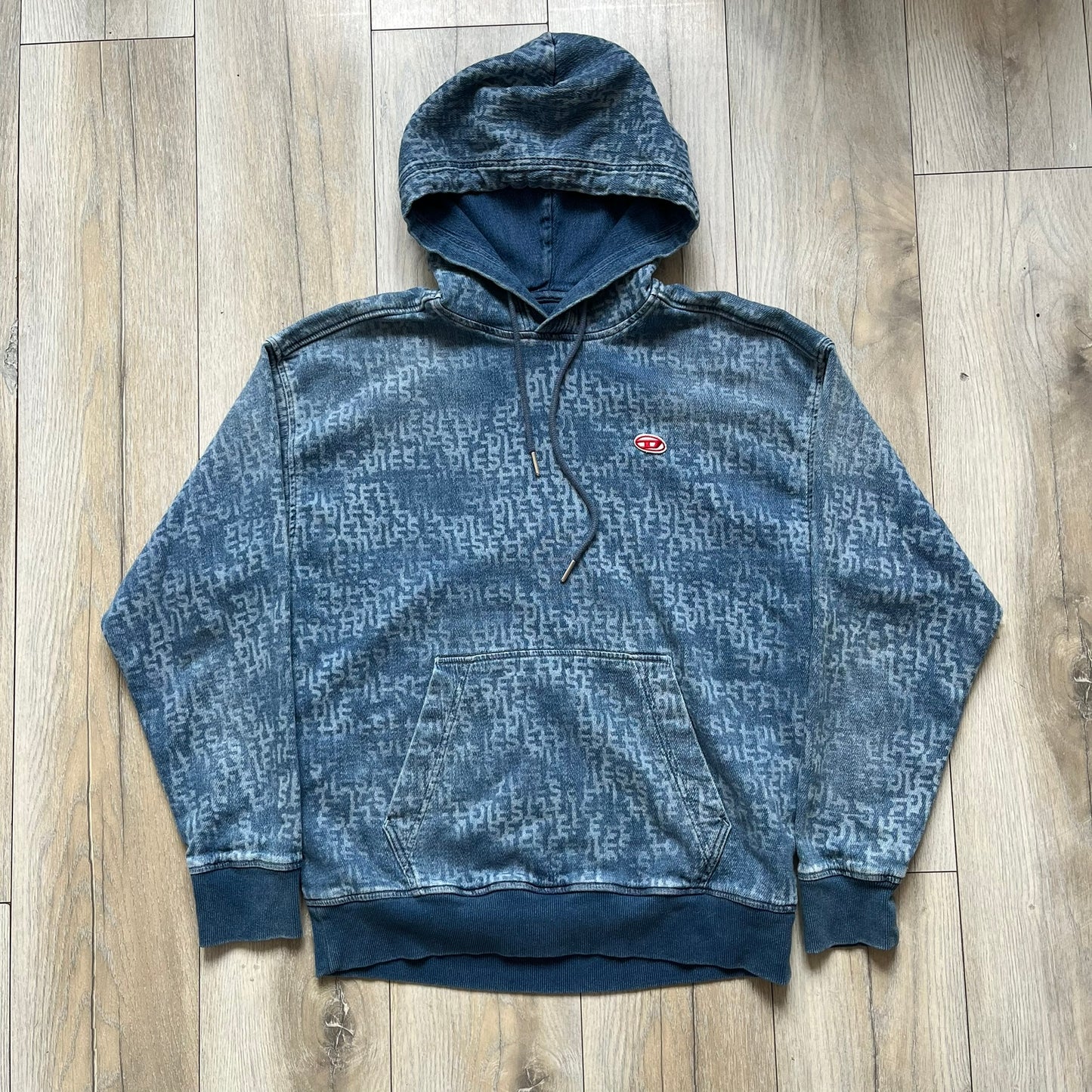 Diesel “Denim” Logo Blue Hoodie is a blue hooded sweatshirt designed with a denim-inspired colorway and washed effect throughout. The fabric features varying light and dark blue shades to mimic the appearance of worn denim, along with an all-over Diesel text pattern in a slightly lighter tone. A small red Diesel patch is placed on the chest, while the cuffs and hem are finished in a contrasting navy color.&nbsp;