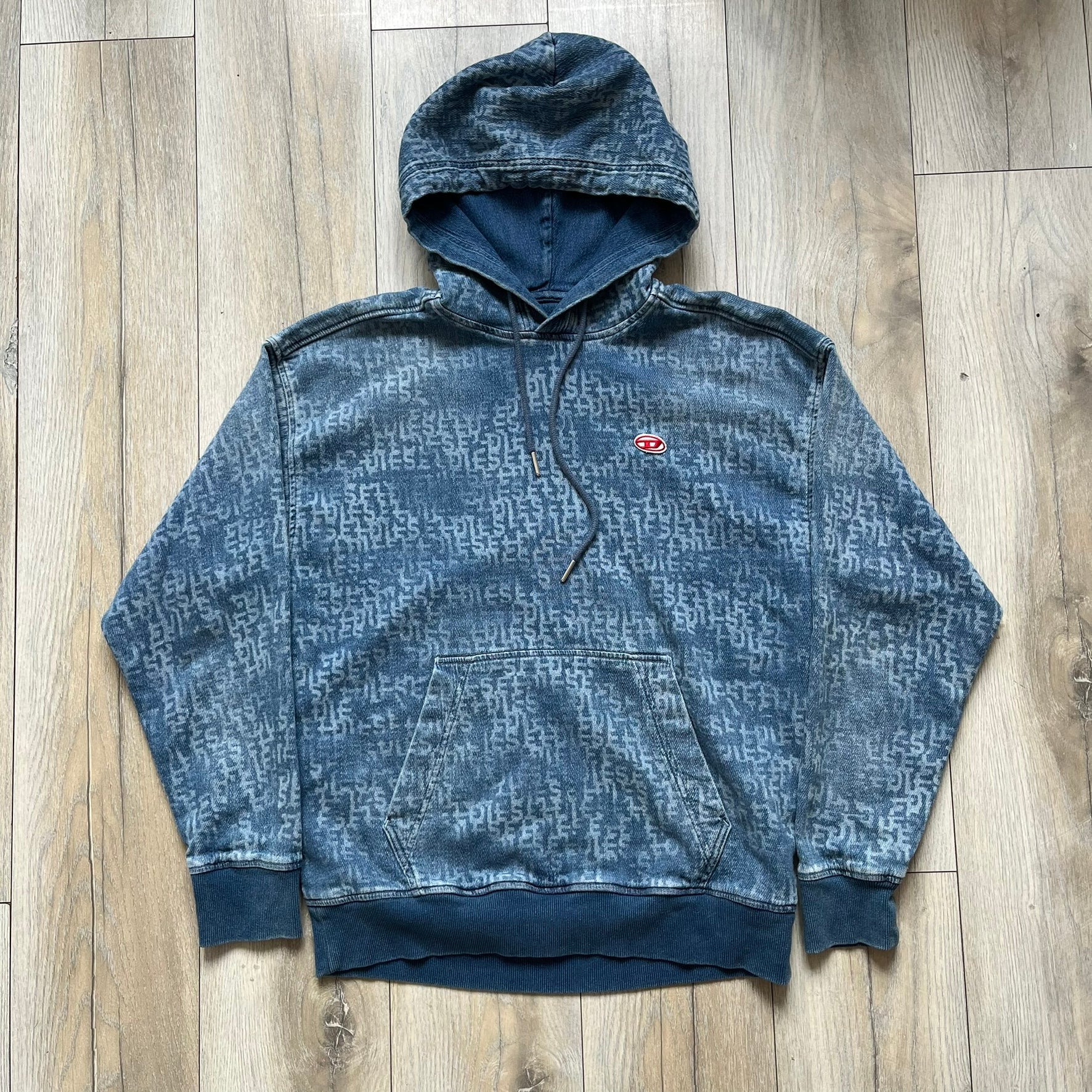 Diesel “Denim” Logo Blue Hoodie is a blue hooded sweatshirt designed with a denim-inspired colorway and washed effect throughout. The fabric features varying light and dark blue shades to mimic the appearance of worn denim, along with an all-over Diesel text pattern in a slightly lighter tone. A small red Diesel patch is placed on the chest, while the cuffs and hem are finished in a contrasting navy color.&nbsp;