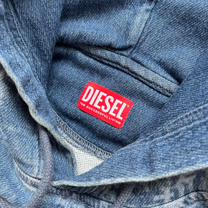 Diesel “Denim” Logo Blue Hoodie is a blue hooded sweatshirt designed with a denim-inspired colorway and washed effect throughout. The fabric features varying light and dark blue shades to mimic the appearance of worn denim, along with an all-over Diesel text pattern in a slightly lighter tone. A small red Diesel patch is placed on the chest, while the cuffs and hem are finished in a contrasting navy color.&nbsp;