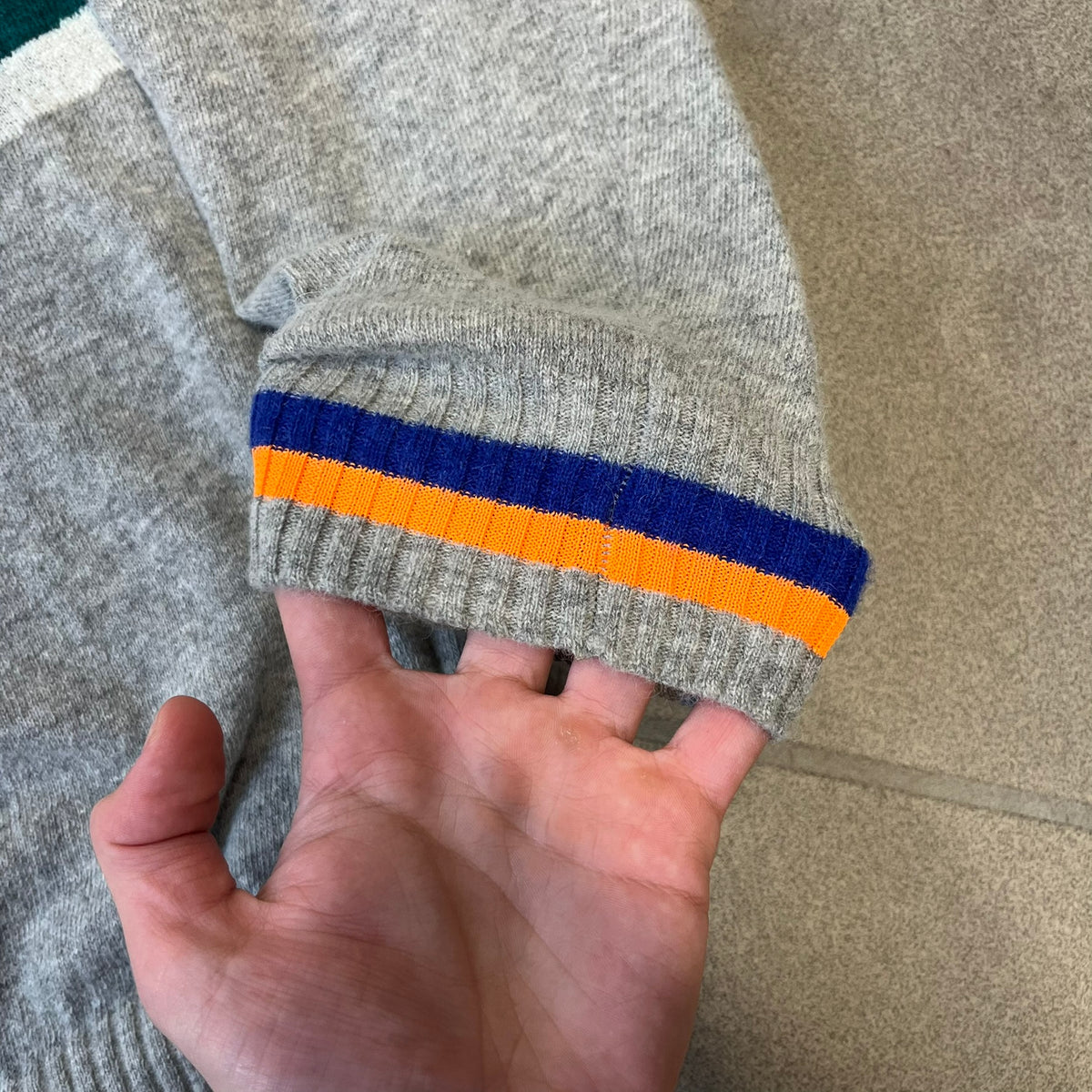 Diesel K-Dino FW19 Gray Pullover is a lightweight gray knit pullover made from alpaca, polyamide, and other materials. The front features a white and black T-rex with a shopping cart, set against multicolor orange, blue, red, and green stripes running behind it.