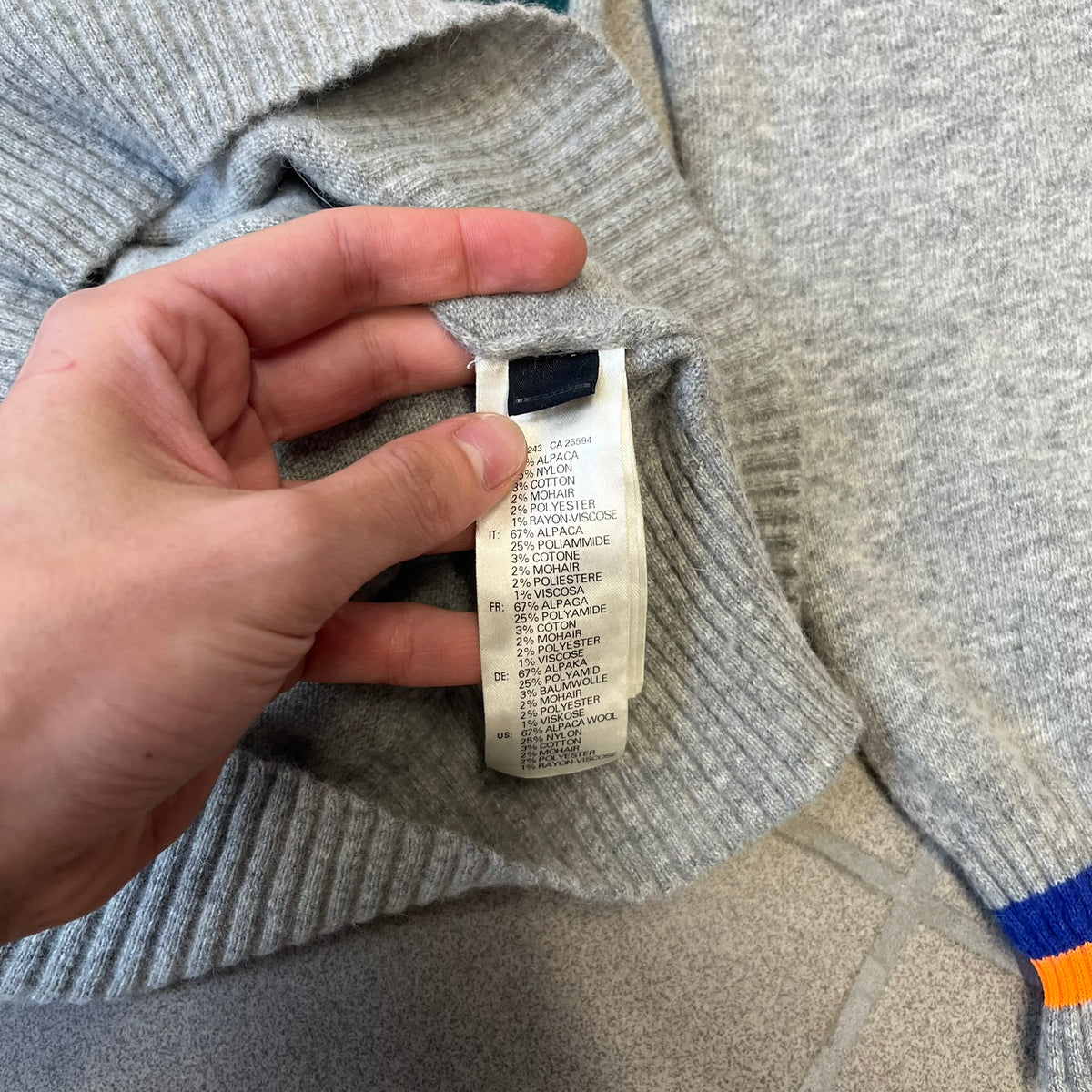 Diesel K-Dino FW19 Gray Pullover is a lightweight gray knit pullover made from alpaca, polyamide, and other materials. The front features a white and black T-rex with a shopping cart, set against multicolor orange, blue, red, and green stripes running behind it.