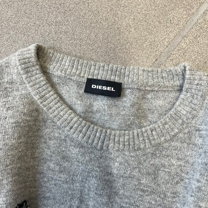 Diesel K-Dino FW19 Gray Pullover is a lightweight gray knit pullover made from alpaca, polyamide, and other materials. The front features a white and black T-rex with a shopping cart, set against multicolor orange, blue, red, and green stripes running behind it.