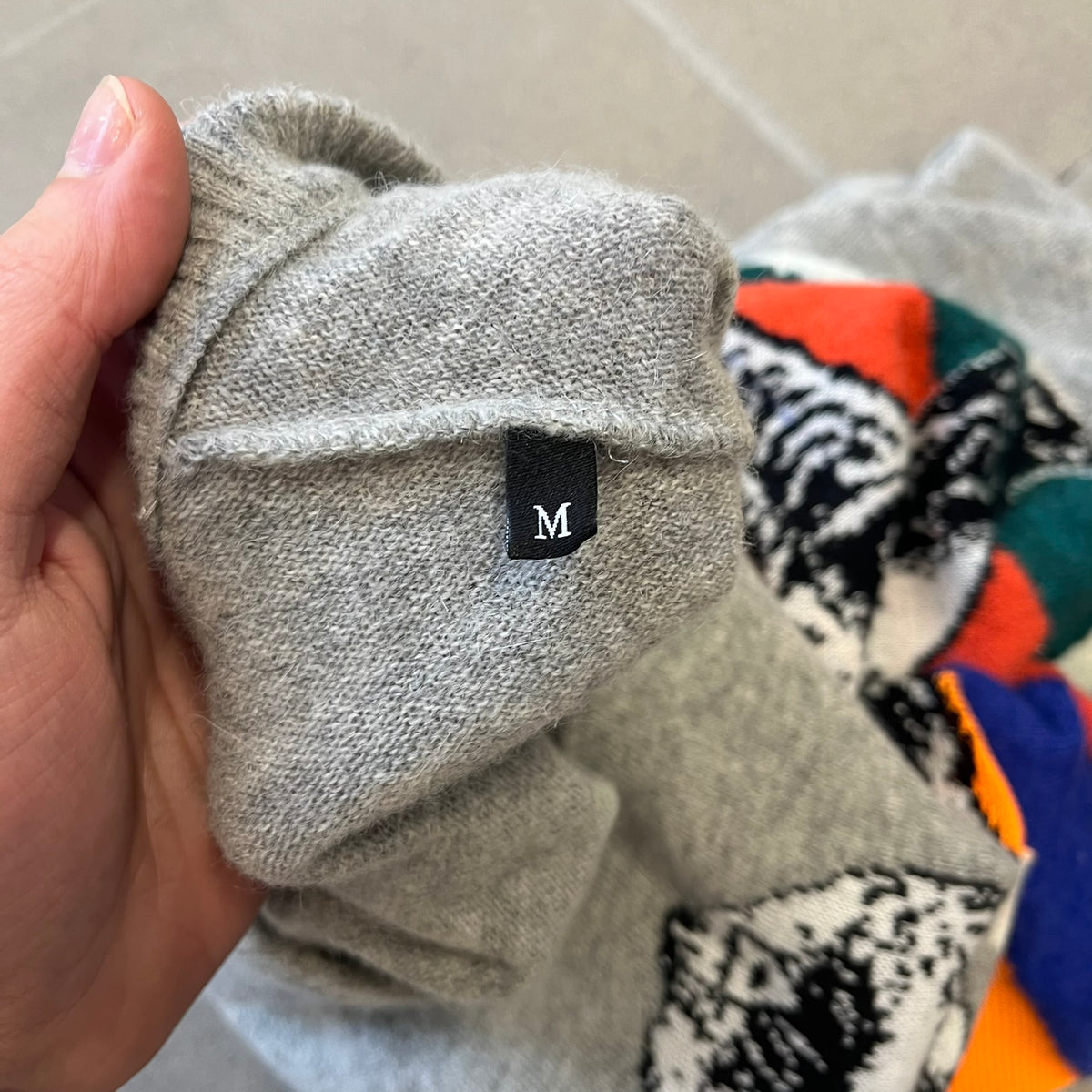 Diesel K-Dino FW19 Gray Pullover is a lightweight gray knit pullover made from alpaca, polyamide, and other materials. The front features a white and black T-rex with a shopping cart, set against multicolor orange, blue, red, and green stripes running behind it.