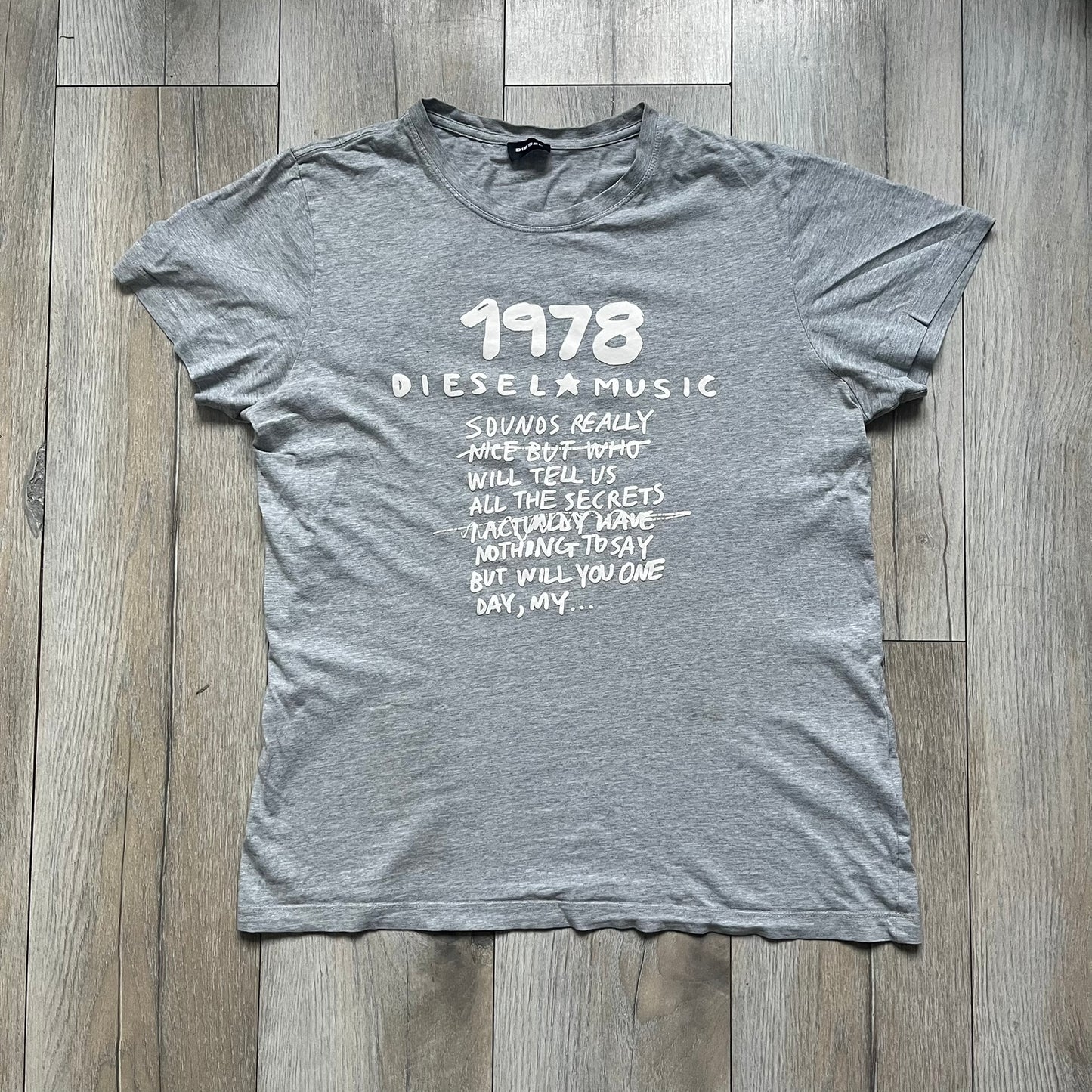 Diesel Music 1978 Gray Tee is a gray T-shirt featuring a large hand-drawn style “Diesel Music 1978” print on the front with additional text below. Made from 100% cotton, it has a crew neckline and short sleeves. The print is white, contrasting with the gray fabric.