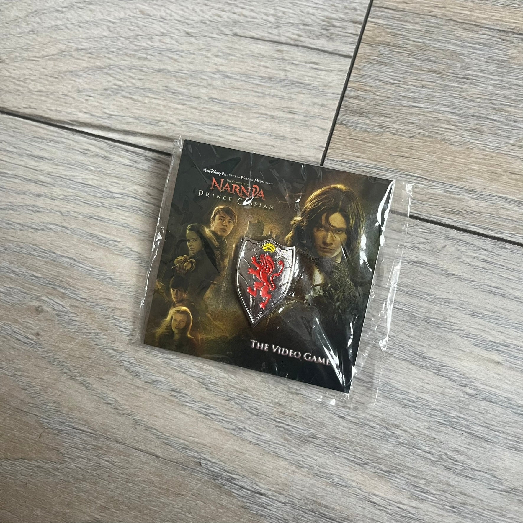 Disney 2008 The Chronicles of Narnia: Prince Caspian Pin is a silver-tone metal pin featuring a red lion with a yellow crown. The pin is attached to a promotional backing card displaying characters from The Chronicles of Narnia: Prince Caspian, the game title, and “The Video Game” text. The reverse side of the card contains additional game information and licensing details.
