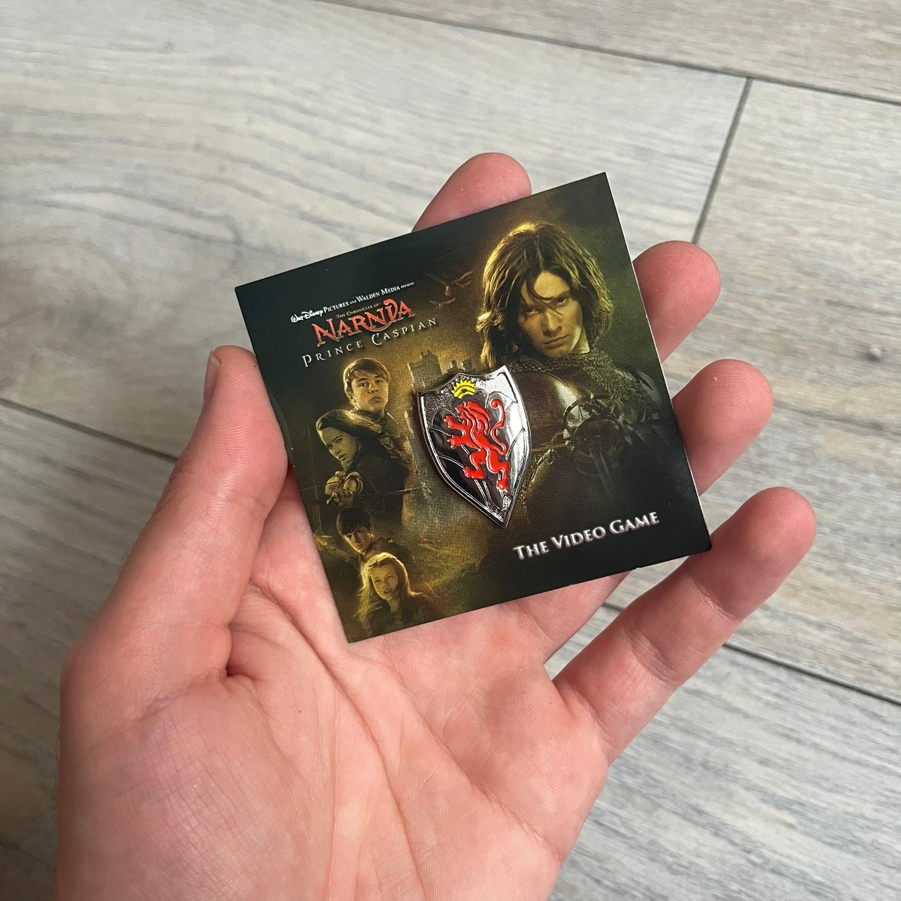 Disney 2008 The Chronicles of Narnia: Prince Caspian Pin is a silver-tone metal pin featuring a red lion with a yellow crown. The pin is attached to a promotional backing card displaying characters from The Chronicles of Narnia: Prince Caspian, the game title, and “The Video Game” text. The reverse side of the card contains additional game information and licensing details.