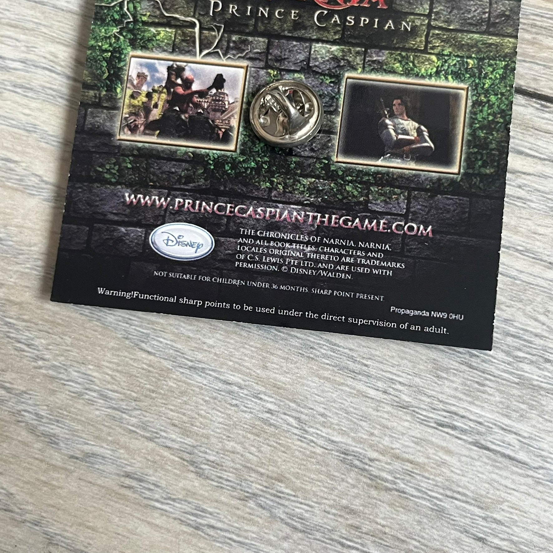 Disney 2008 The Chronicles of Narnia: Prince Caspian Pin is a silver-tone metal pin featuring a red lion with a yellow crown. The pin is attached to a promotional backing card displaying characters from The Chronicles of Narnia: Prince Caspian, the game title, and “The Video Game” text. The reverse side of the card contains additional game information and licensing details.