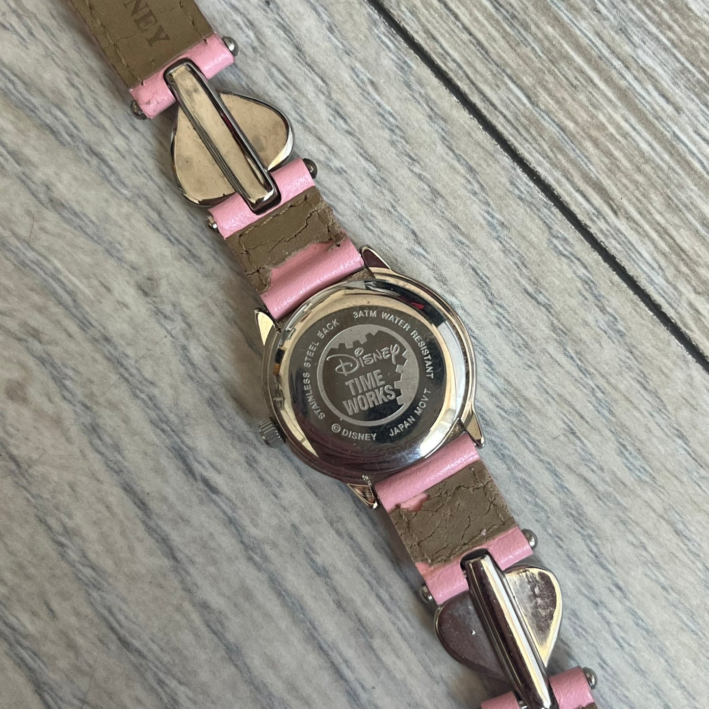 Disney Mickey Mouse 1990s Pink Analog Watch features a pink leather bracelet with metal heart details connecting sections of the strap. It has a silver-tone case and a silver dial with matching silver hour markers and hands. The center of the dial features a Mickey Mouse graphic with a small Disney logo beside it. The back includes a Time Works logo and Japan movement marking. The watch is sized for smaller wrists.
