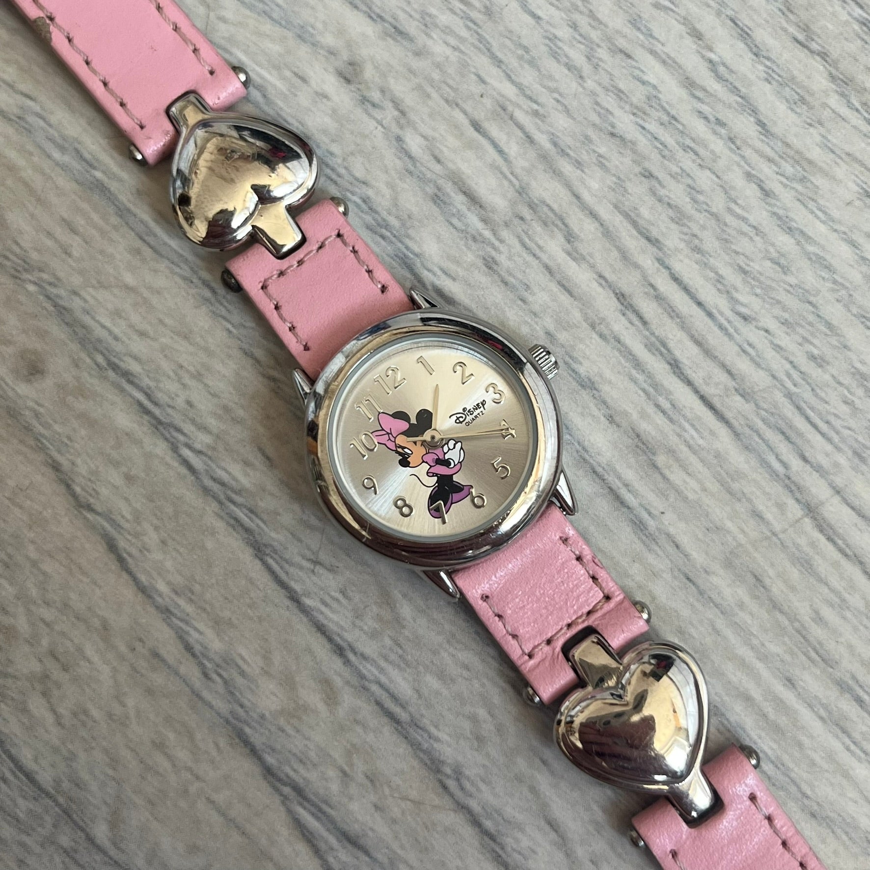 Disney Mickey Mouse 1990s Pink Analog Watch features a pink leather bracelet with metal heart details connecting sections of the strap. It has a silver-tone case and a silver dial with matching silver hour markers and hands. The center of the dial features a Mickey Mouse graphic with a small Disney logo beside it. The back includes a Time Works logo and Japan movement marking. The watch is sized for smaller wrists.
