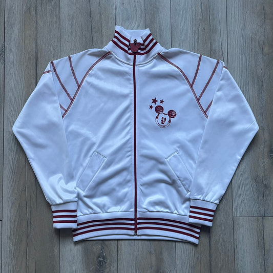 White 1990s Disney track jacket with red contrast details throughout. Features red stitching and accent lines, a red zip closure with a large red metal Mickey Mouse zipper pull, and a small Mickey Mouse face with red stars on the chest. The back has a large Mickey Mouse graphic with red stars and oversized “Tokyo” text printed above.