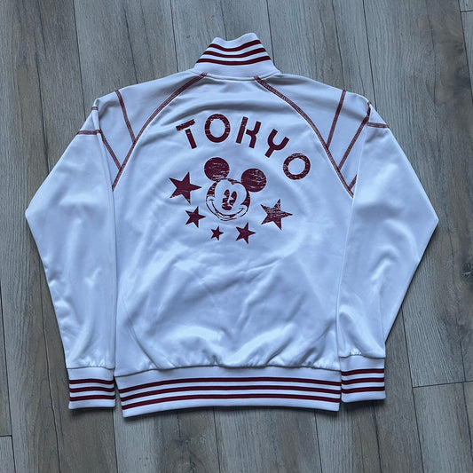 White 1990s Disney track jacket with red contrast details throughout. Features red stitching and accent lines, a red zip closure with a large red metal Mickey Mouse zipper pull, and a small Mickey Mouse face with red stars on the chest. The back has a large Mickey Mouse graphic with red stars and oversized “Tokyo” text printed above.
