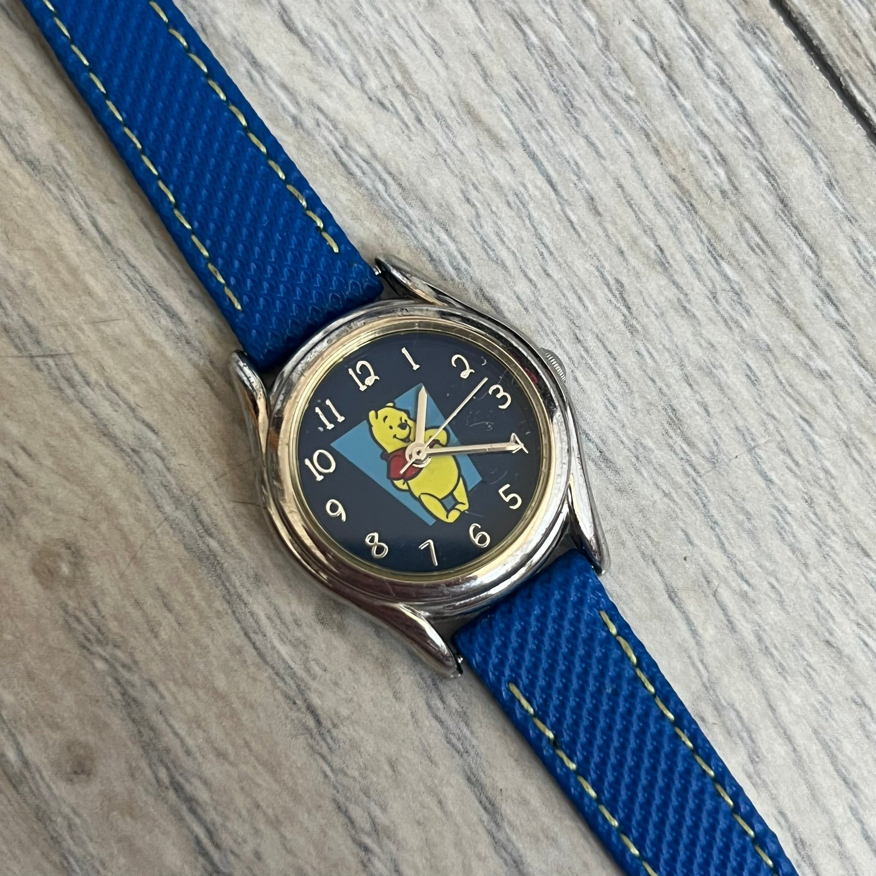 Disney Winnie-the-Pooh 1990s Analog Watch features blue leather straps, a gold-tone case, and a navy dial. The dial displays gold hour markers with Winnie the Pooh illustrated in the center. The case back is marked with official Disney licensing and indicates it was made exclusively for Disney. This watch is sized for smaller wrists.
