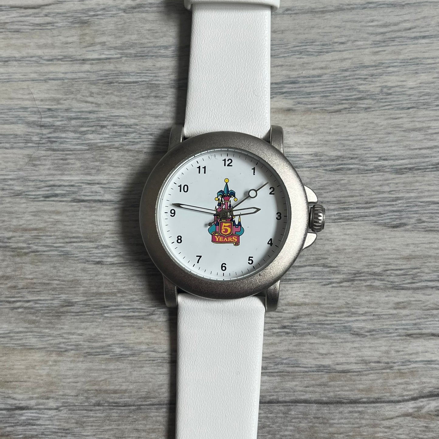 Disneyland Paris 1997 “5 Years Anniversary” Analog Watch features a silver-tone metal case, white dial, and white leather straps. The dial displays a colorful Disney castle graphic with “5 Years” text commemorating the anniversary of the Paris location. The case back is marked “Disneyland Paris Avronel” and indicates production by Avronel for Disney. The watch is rated for 3 ATM water resistance.