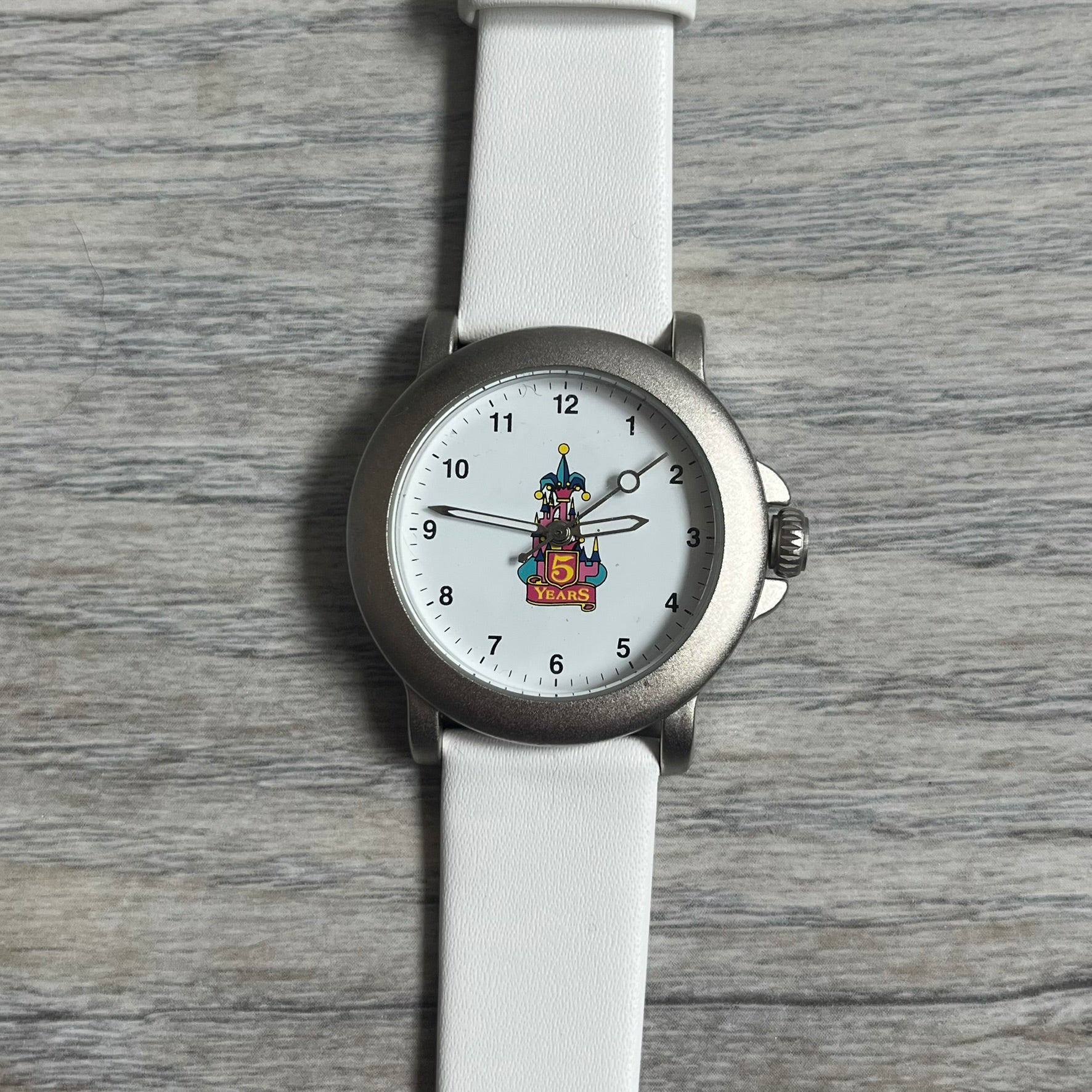 Disneyland Paris 1997 “5 Years Anniversary” Analog Watch features a silver-tone metal case, white dial, and white leather straps. The dial displays a colorful Disney castle graphic with “5 Years” text commemorating the anniversary of the Paris location. The case back is marked “Disneyland Paris Avronel” and indicates production by Avronel for Disney. The watch is rated for 3 ATM water resistance.