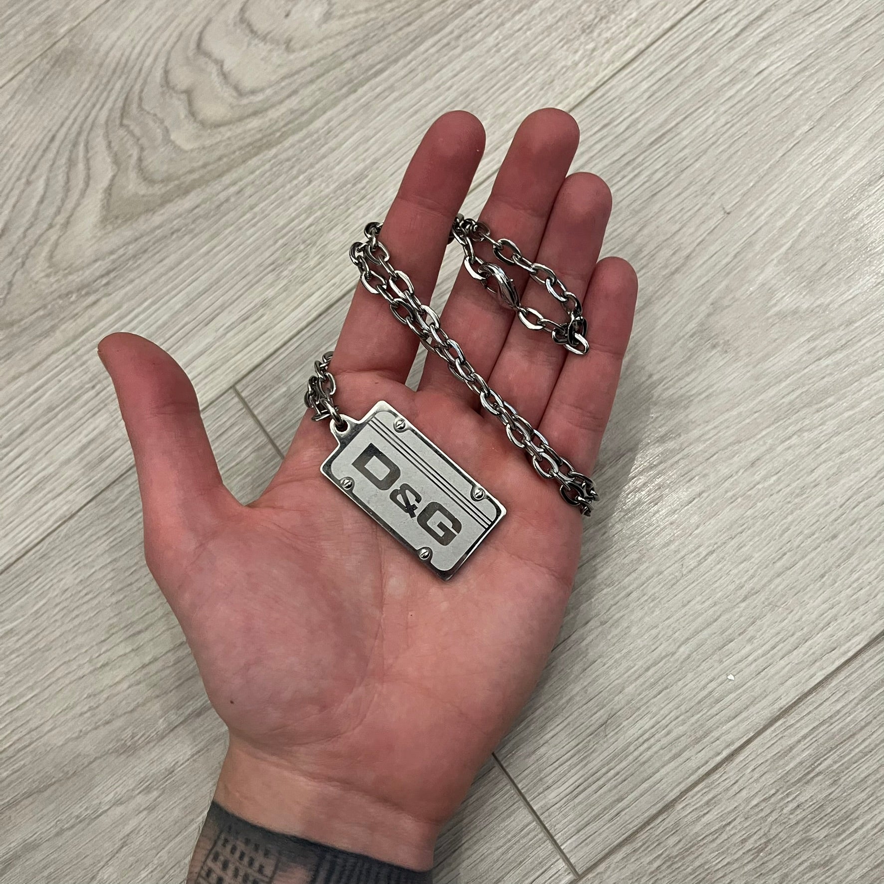 Dolce &amp; Gabbana Propaganda Dog Tag Necklace, featuring a silver stainless steel square tag with four visible screws and the D&amp;G logo engraved on the front. The tag hangs from a durable cable chain, combining sturdy construction with clean, industrial-inspired detailing.