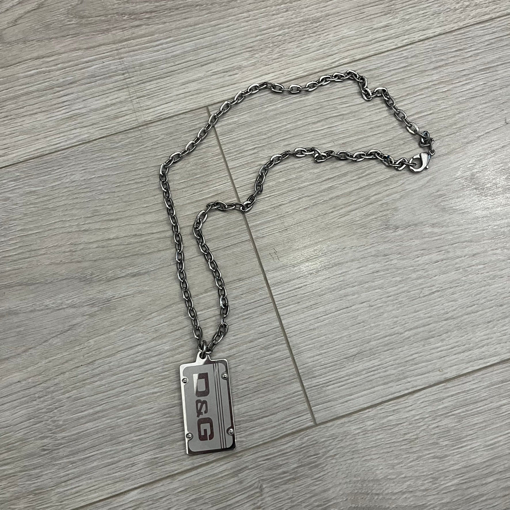 Dolce &amp; Gabbana Propaganda Dog Tag Necklace, featuring a silver stainless steel square tag with four visible screws and the D&amp;G logo engraved on the front. The tag hangs from a durable cable chain, combining sturdy construction with clean, industrial-inspired detailing.