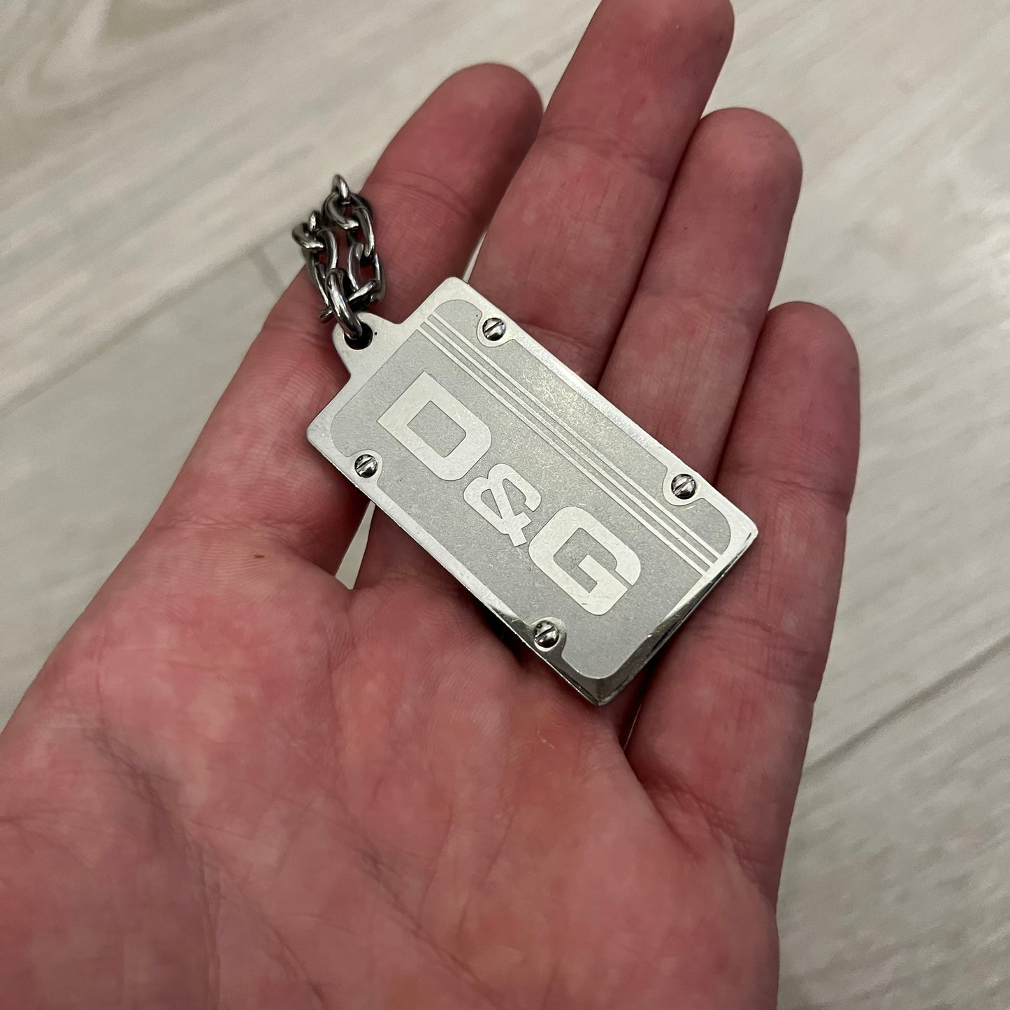 Dolce &amp; Gabbana Propaganda Dog Tag Necklace, featuring a silver stainless steel square tag with four visible screws and the D&amp;G logo engraved on the front. The tag hangs from a durable cable chain, combining sturdy construction with clean, industrial-inspired detailing.