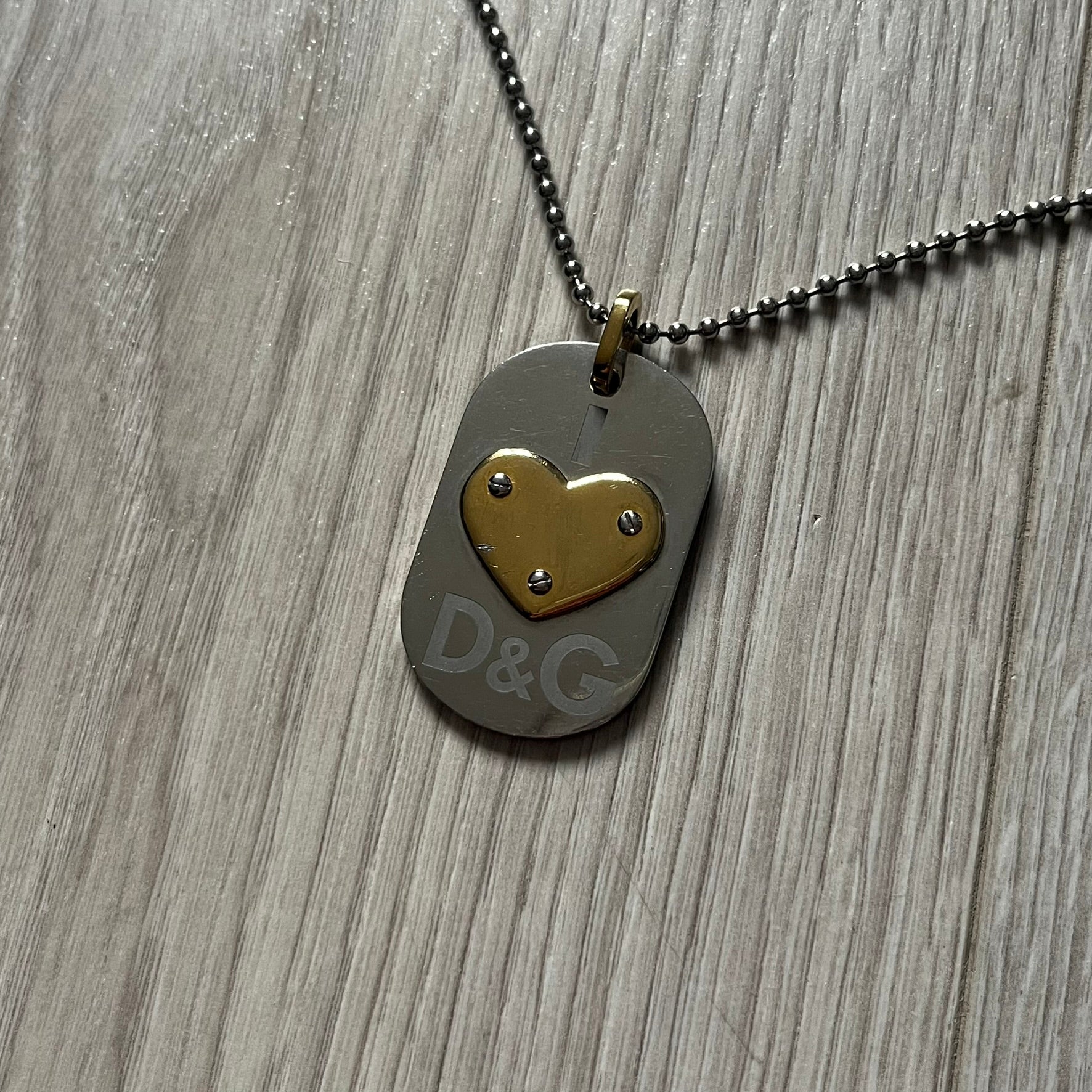 Dolce &amp; Gabbana "I ❤️ D&G" Gold Dog Tag Necklace with a silver stainless steel ball chain, featuring a dog tag engraved with a light "I", a gold metal heart with 3 visible screws, and "D&G" text below, forming the "I ❤️ D&G" design. A playful and collectible vintage piece.