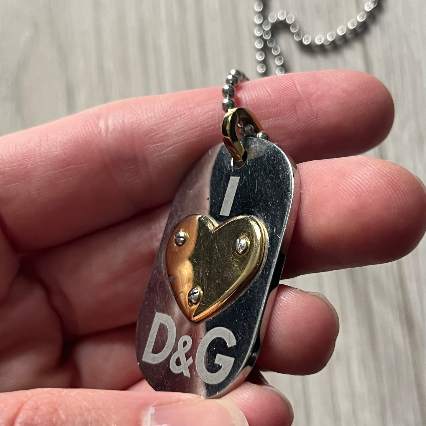 Dolce & Gabbana "I ❤️ D&G" Gold Dog Tag Necklace with a silver stainless steel ball chain, featuring a dog tag engraved with a light "I", a gold metal heart with 3 visible screws, and "D&G" text below, forming the "I ❤️ D&G" design. A playful and collectible vintage piece.