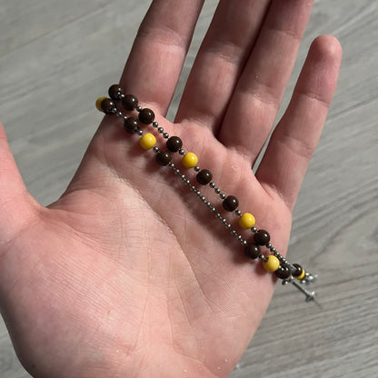 Dolce & Gabbana Pearl Brown Rosary Necklace from the 2000s era. The necklace features a silver-tone metal cross with a D&G logo, connected via a black leather link to a metal chain with alternating brown and yellow pearl-style beads. Rosary-style construction with mixed materials including metal, leather, and resin pearls. Comes complete with the original box.