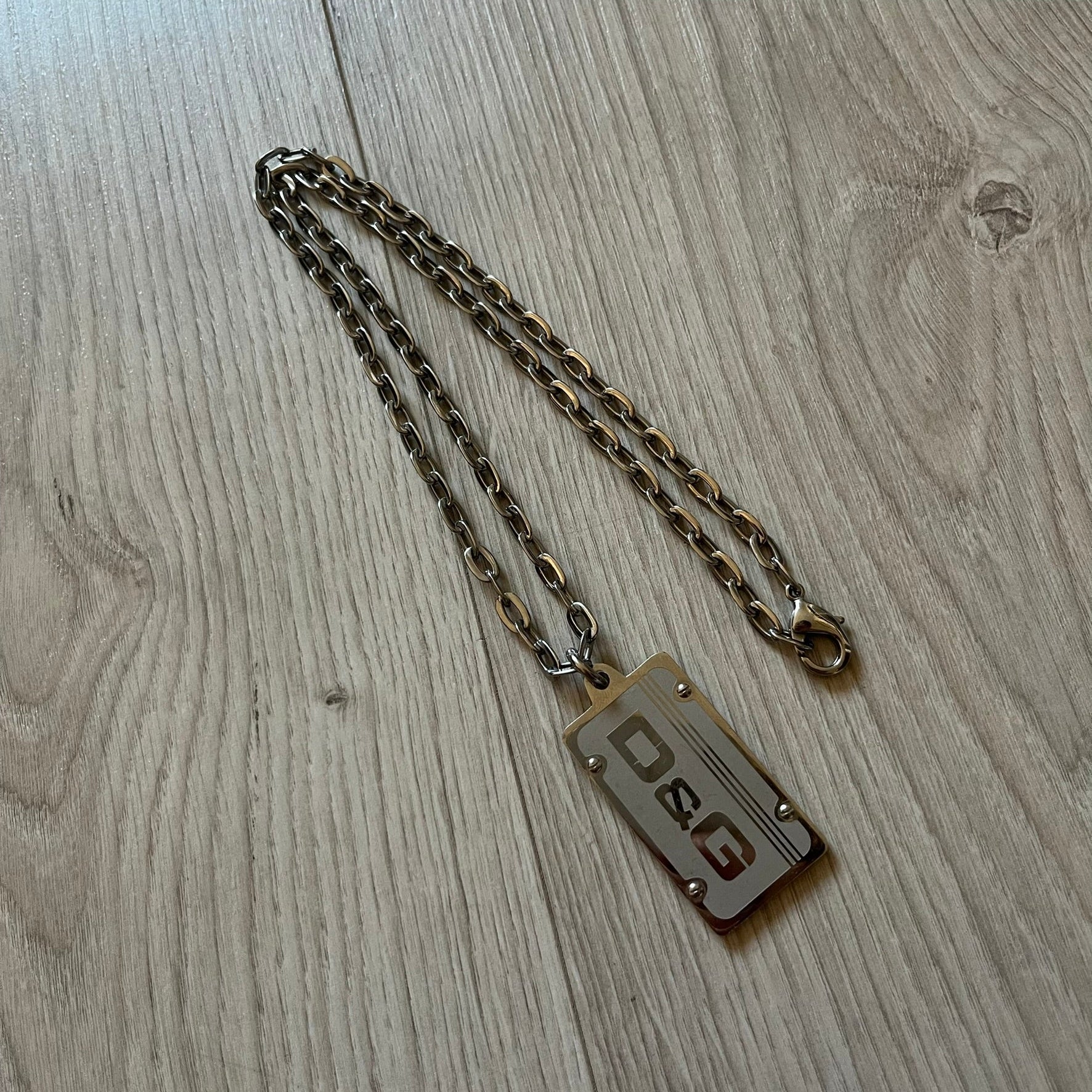 Dolce &amp; Gabbana Propaganda Dog Tag Necklace, featuring a silver stainless steel square tag with four visible screws and the D&amp;G logo engraved on the front. The tag hangs from a durable cable chain, combining sturdy construction with clean, industrial-inspired detailing.