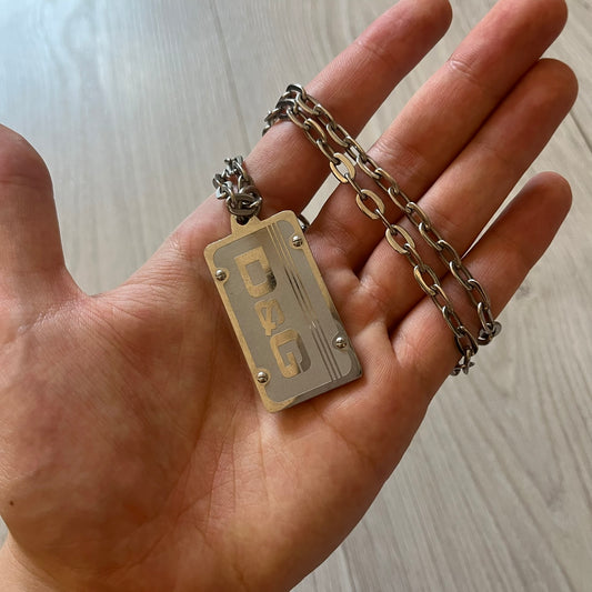 Dolce &amp; Gabbana Propaganda Dog Tag Necklace, featuring a silver stainless steel square tag with four visible screws and the D&amp;G logo engraved on the front. The tag hangs from a durable cable chain, combining sturdy construction with clean, industrial-inspired detailing.
