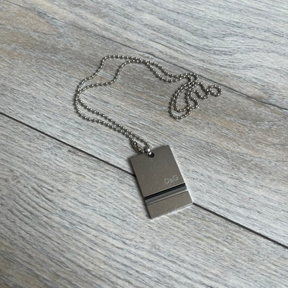 Dolce & Gabbana 2000s 3 Lines Dog Tag Necklace is a metal dog tag pendant featuring an engraved D&G logo positioned slightly to the right on the front. Below it are three horizontal lines in black, grey, and white running across the pendant. The back is plain with four visible screws, and it comes with a standard steel ball chain.
