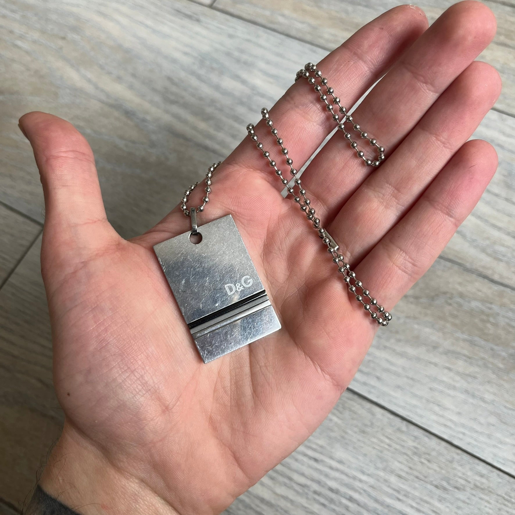 Dolce & Gabbana 2000s 3 Lines Dog Tag Necklace is a metal dog tag pendant featuring an engraved D&G logo positioned slightly to the right on the front. Below it are three horizontal lines in black, grey, and white running across the pendant. The back is plain with four visible screws, and it comes with a standard steel ball chain.
