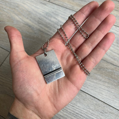 Dolce & Gabbana 2000s 3 Lines Dog Tag Necklace is a metal dog tag pendant featuring an engraved D&G logo positioned slightly to the right on the front. Below it are three horizontal lines in black, grey, and white running across the pendant. The back is plain with four visible screws, and it comes with a standard steel ball chain.
