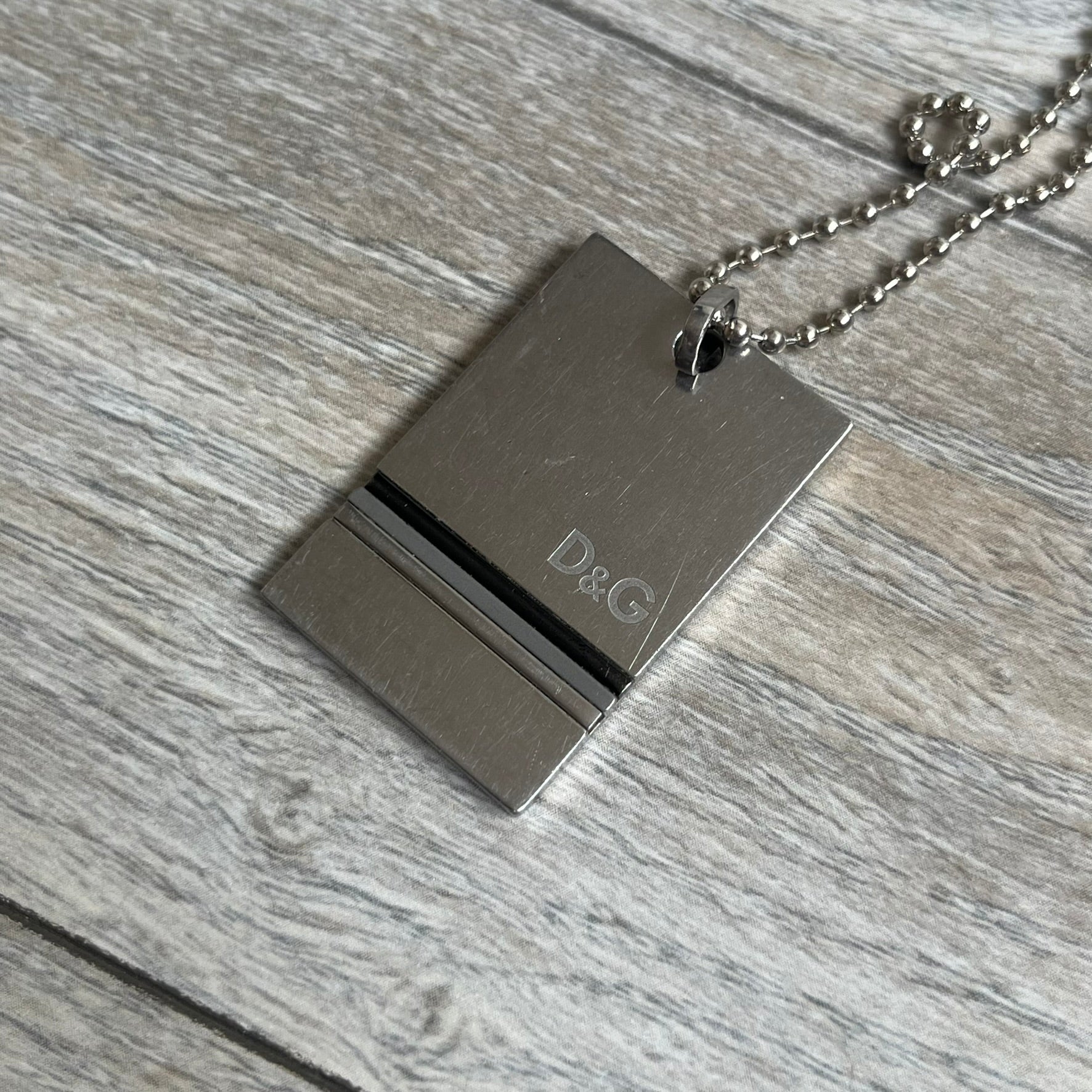 Dolce & Gabbana 2000s 3 Lines Dog Tag Necklace is a metal dog tag pendant featuring an engraved D&G logo positioned slightly to the right on the front. Below it are three horizontal lines in black, grey, and white running across the pendant. The back is plain with four visible screws, and it comes with a standard steel ball chain.
