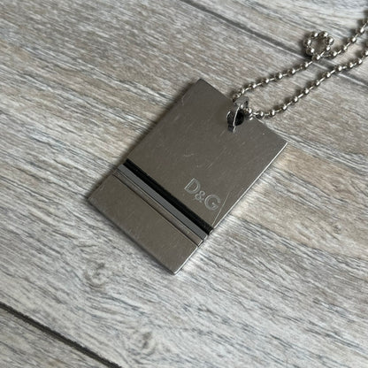 Dolce & Gabbana 2000s 3 Lines Dog Tag Necklace is a metal dog tag pendant featuring an engraved D&G logo positioned slightly to the right on the front. Below it are three horizontal lines in black, grey, and white running across the pendant. The back is plain with four visible screws, and it comes with a standard steel ball chain.
