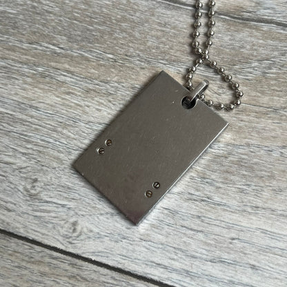 Dolce & Gabbana 2000s 3 Lines Dog Tag Necklace is a metal dog tag pendant featuring an engraved D&G logo positioned slightly to the right on the front. Below it are three horizontal lines in black, grey, and white running across the pendant. The back is plain with four visible screws, and it comes with a standard steel ball chain.
