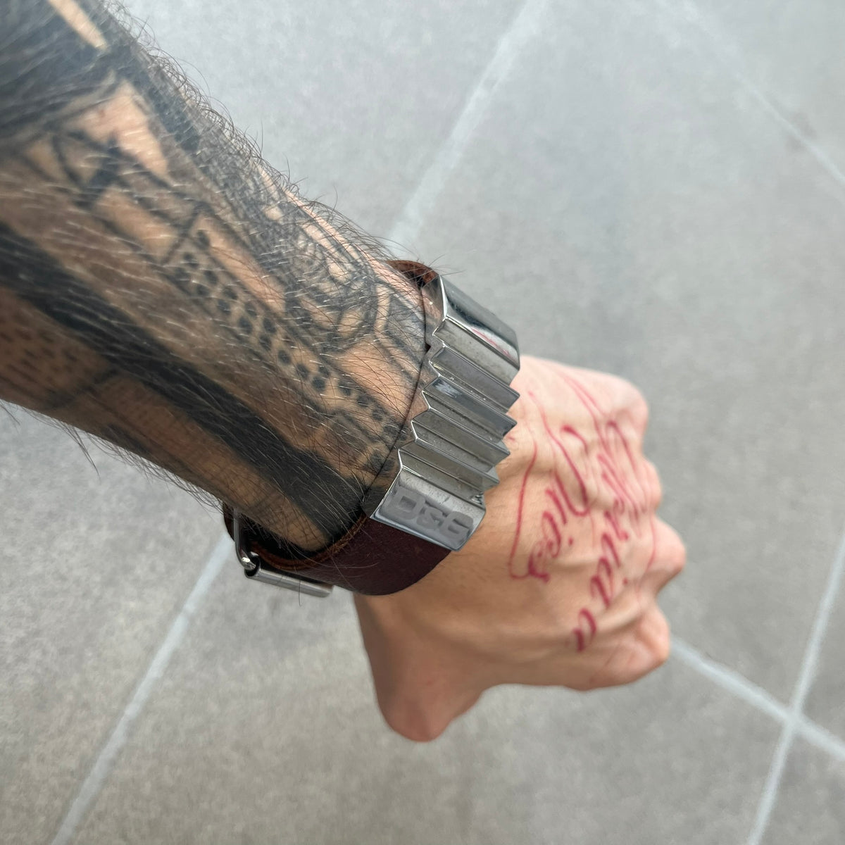 Dolce & Gabbana 2000's Brown Leather Bracelet features brown leather straps with an adjustable fit and a stainless steel central plaque. The plaque has a raised wavy texture on the front and a D&G logo underneath, while the leather straps have small D&G and “Genuine Leather” tags.
