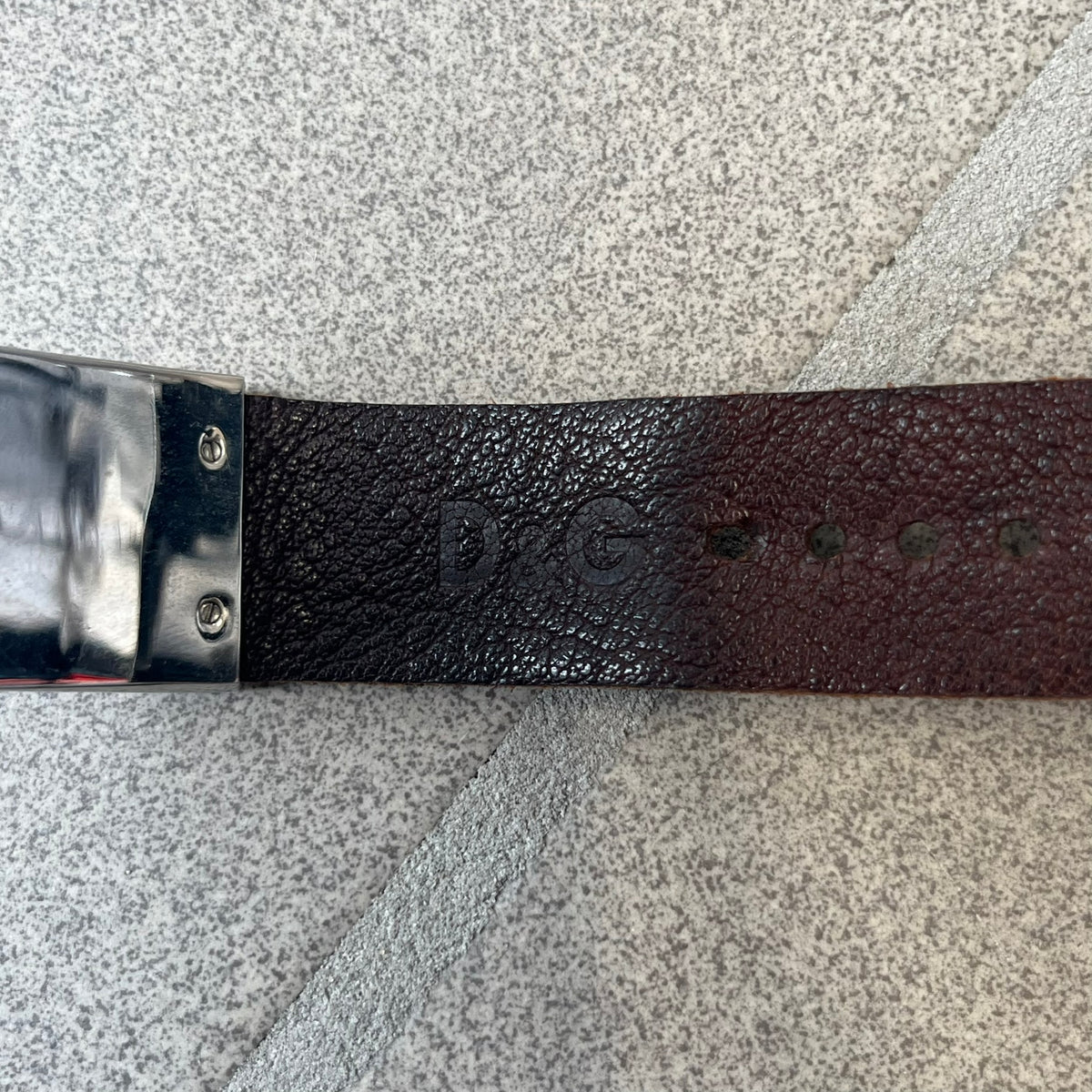 Dolce & Gabbana 2000's Brown Leather Bracelet features brown leather straps with an adjustable fit and a stainless steel central plaque. The plaque has a raised wavy texture on the front and a D&G logo underneath, while the leather straps have small D&G and “Genuine Leather” tags.
